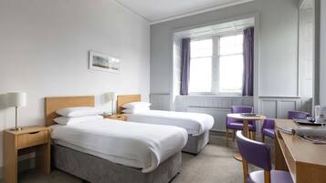 Habitación de hotel con dos camas individuales, un escritorio con silla, una mesita y una ventana con cortinas.