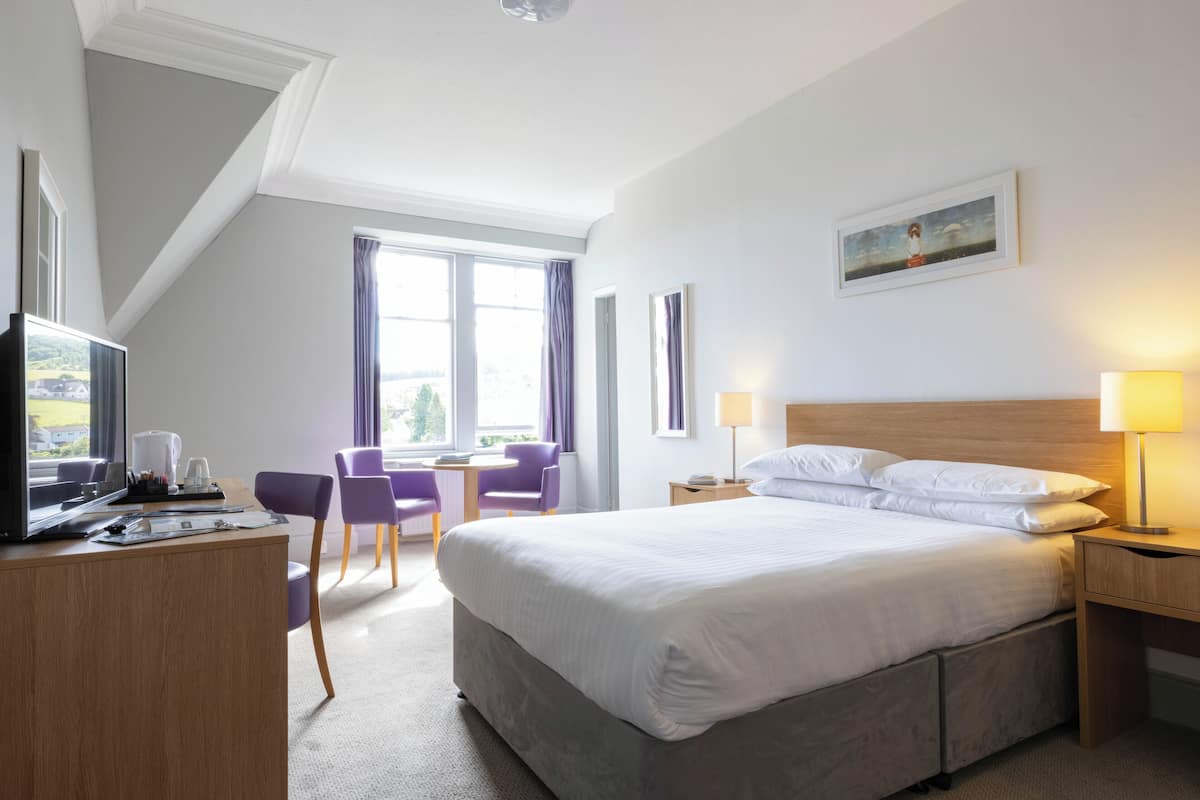 Quarto de hotel com uma cama grande, uma escrivaninha com computador, uma cadeira e uma janela com cortinas.