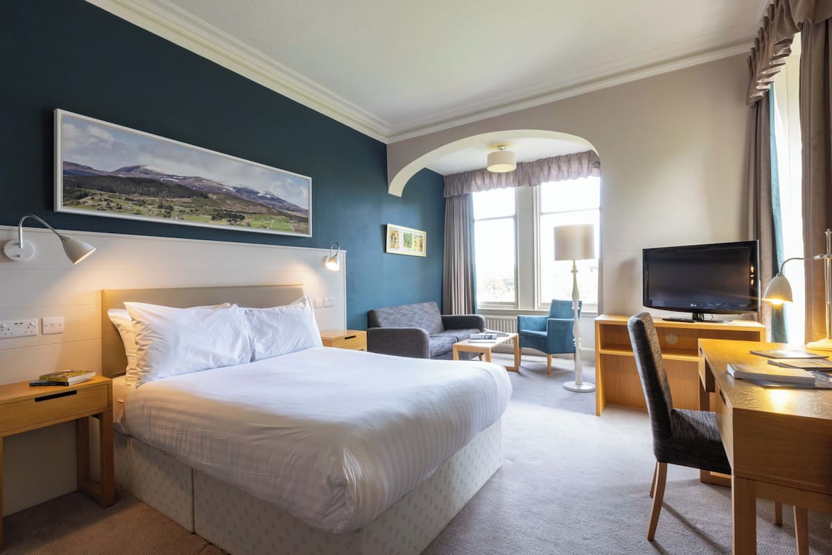 Quarto de hotel com uma cama grande, uma mesa com computador, uma televisão e um espaço de estar com sofá e poltronas.
