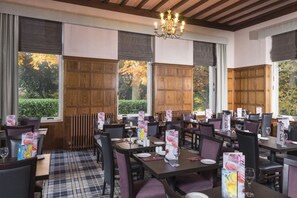 Un restaurante con paredes de madera, amplias ventanas con vistas a árboles y una araña. Las mesas están preparadas con menús y copas de vino.