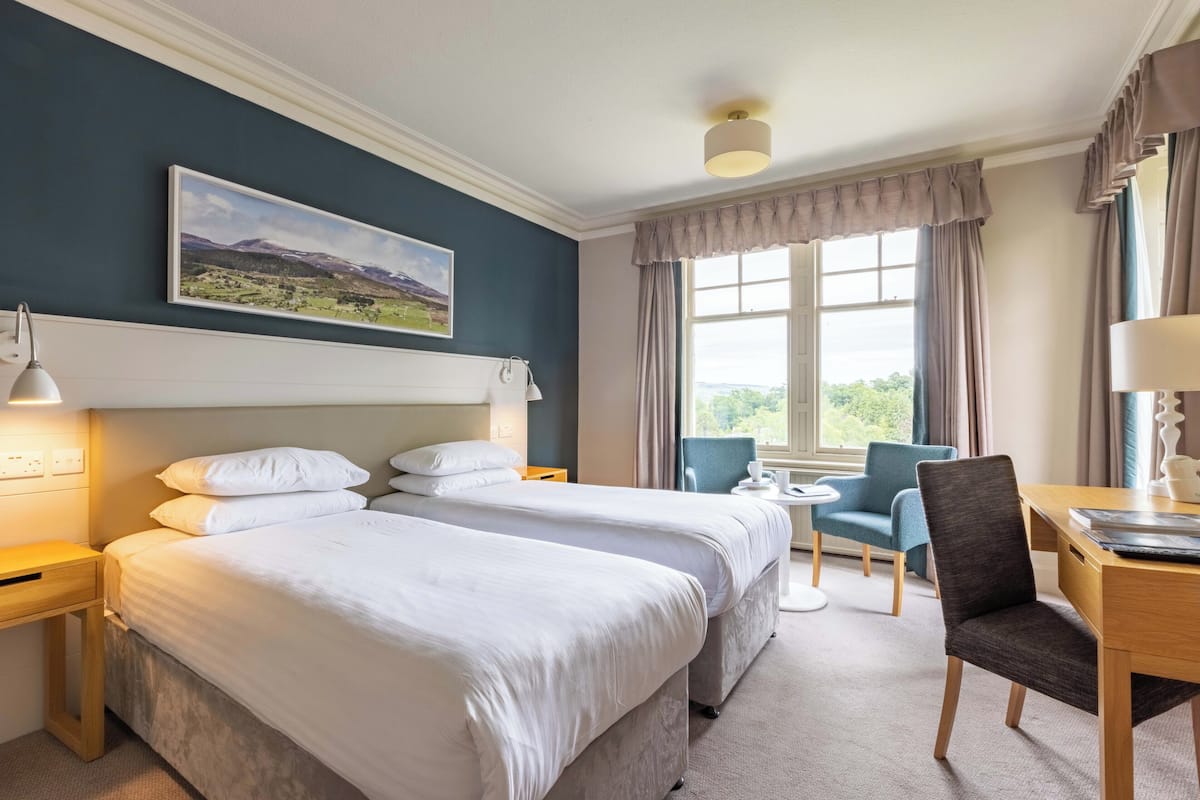 Quarto de hotel com duas camas, uma escrivaninha, uma cadeira, uma janela com cortinas e um quadro na parede.