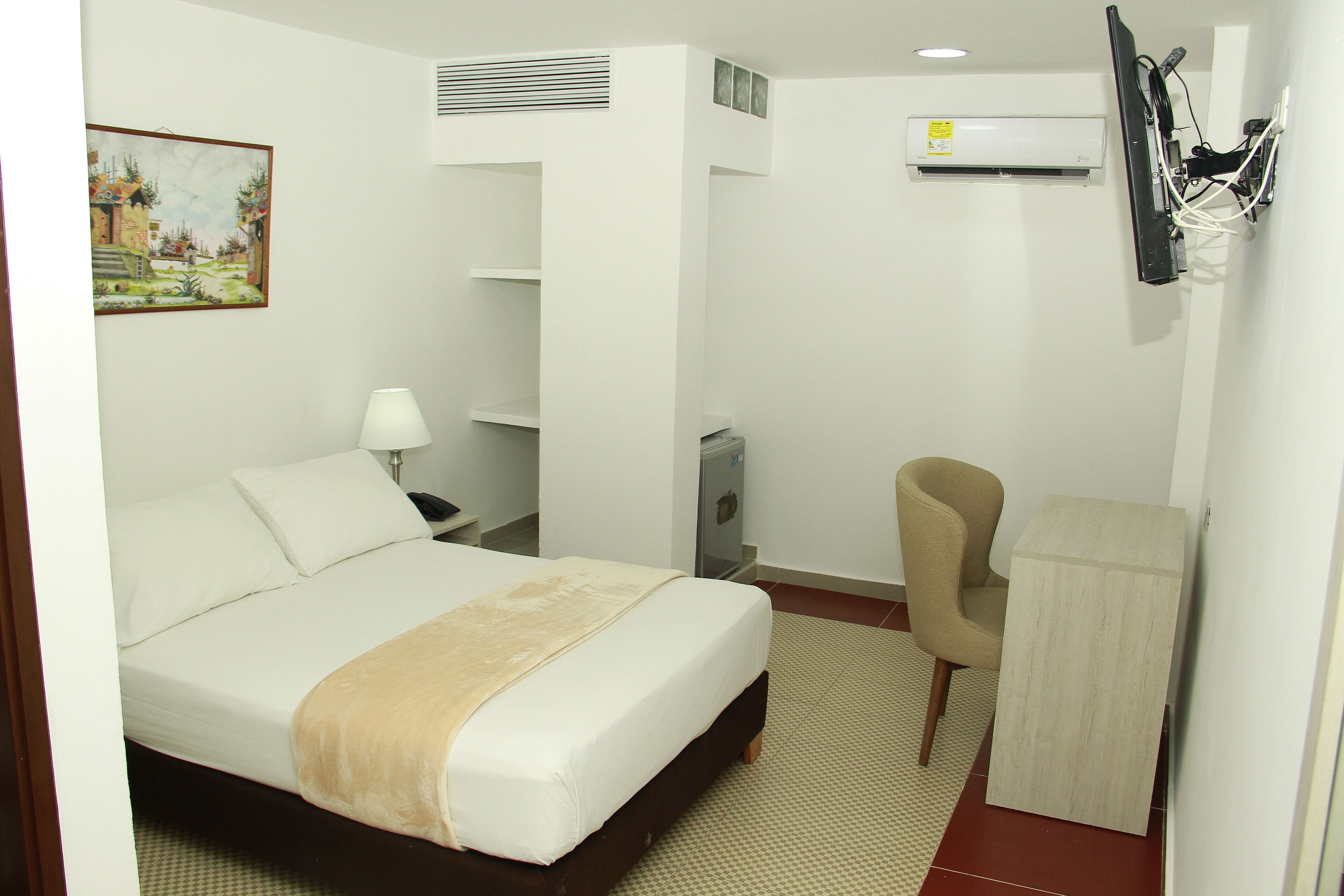 A hotel room with a bed, a chair, a desk, an air conditioner, and a painting on the wall.