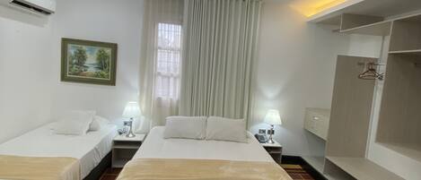 A hotel room with two single beds, a large bed, a chandelier, a painting, a window with curtains, an air conditioning unit, a wardrobe, and a tiled floor.