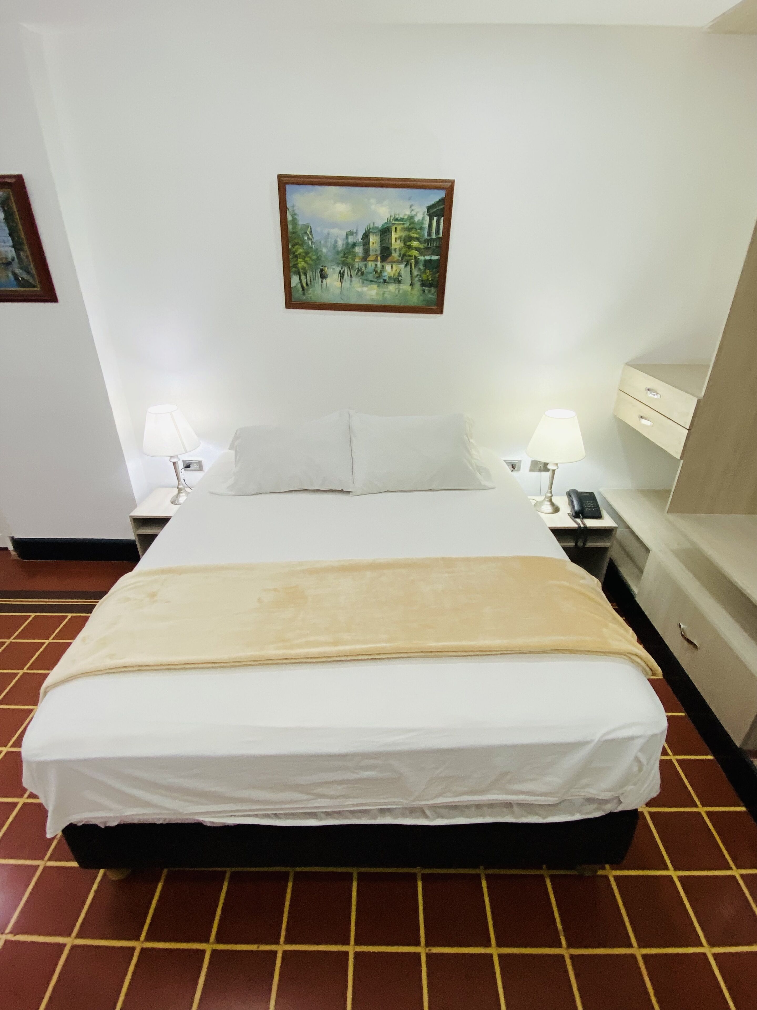 A hotel room with a bed, two bedside lamps, a nightstand, a telephone, a small table, and a painting on the wall.