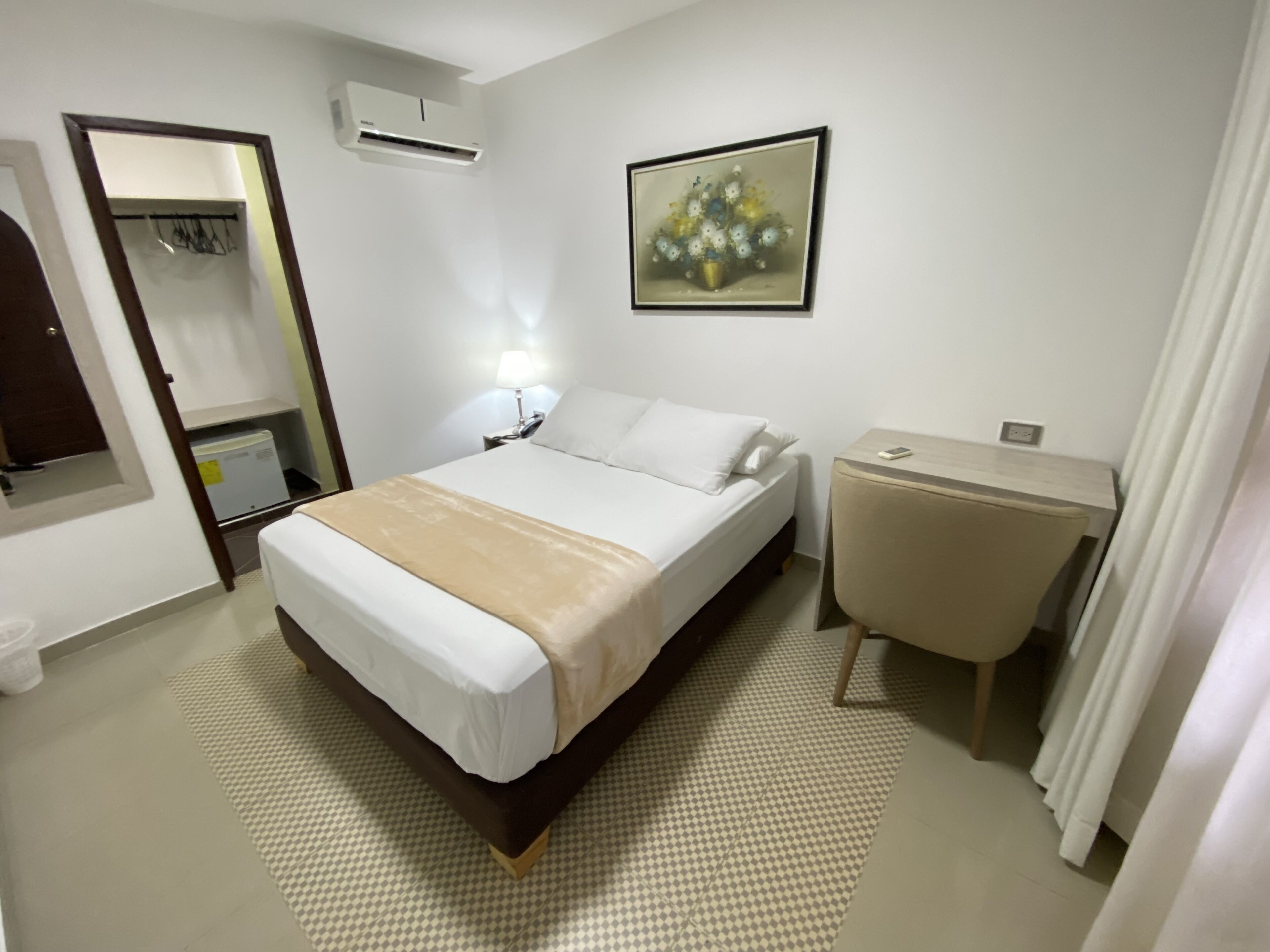 Habitación de hotel con cama, mesita de noche, lámpara, unidad de aire acondicionado y un cuadro en la pared.