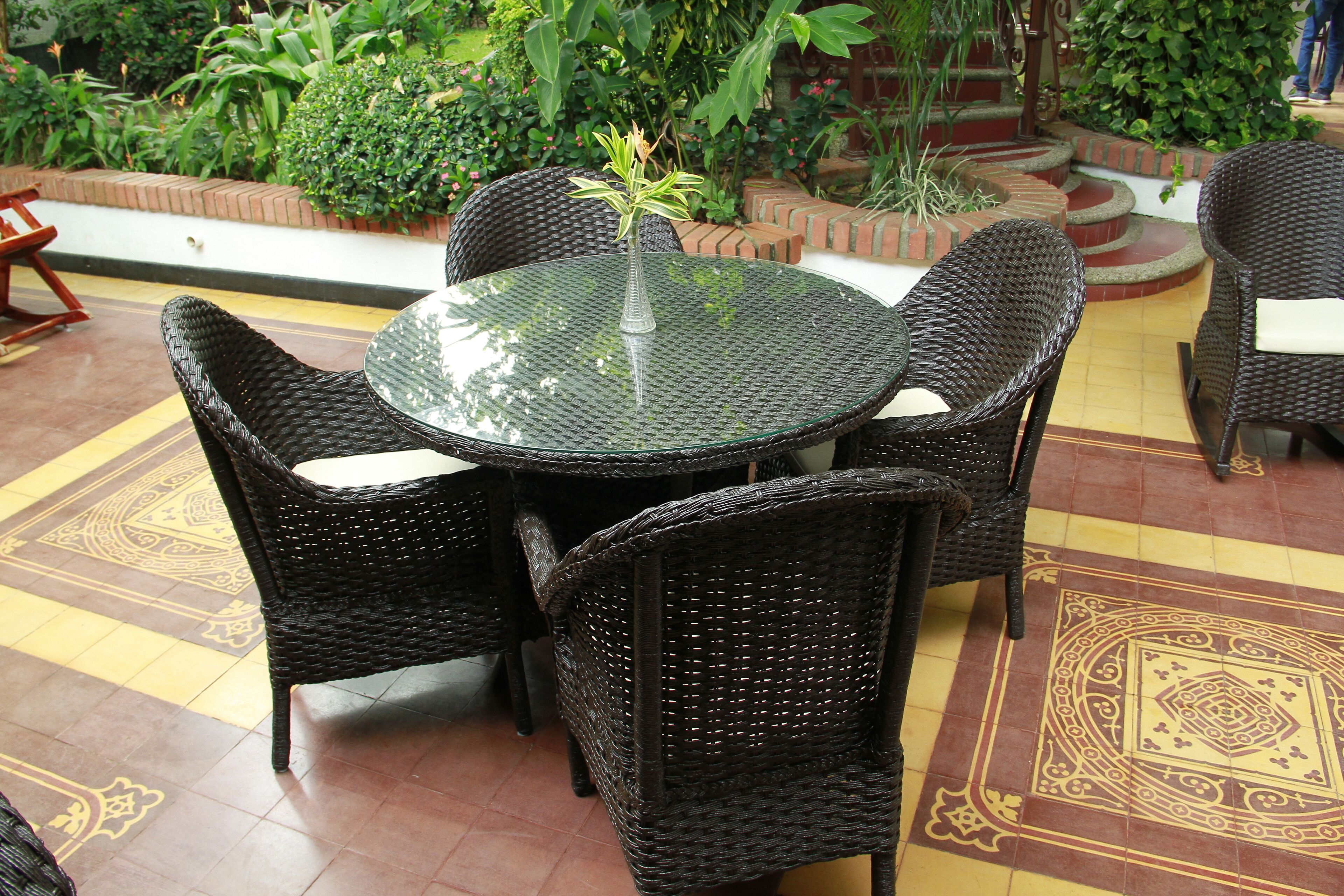 Una mesa redonda de vidrio con sillas de mimbre, rodeada por una jardinera de ladrillo y follaje.