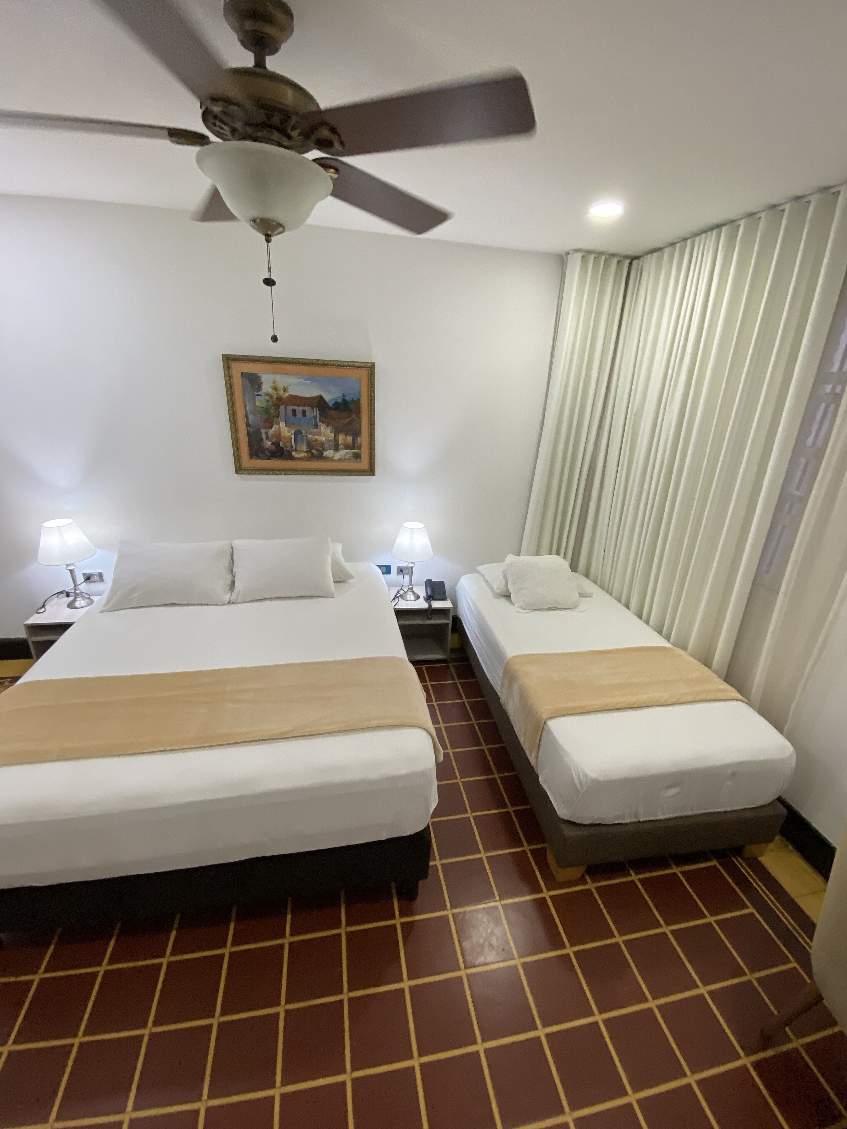 A hotel room with two beds, a ceiling fan, a painting, and a window with curtains.