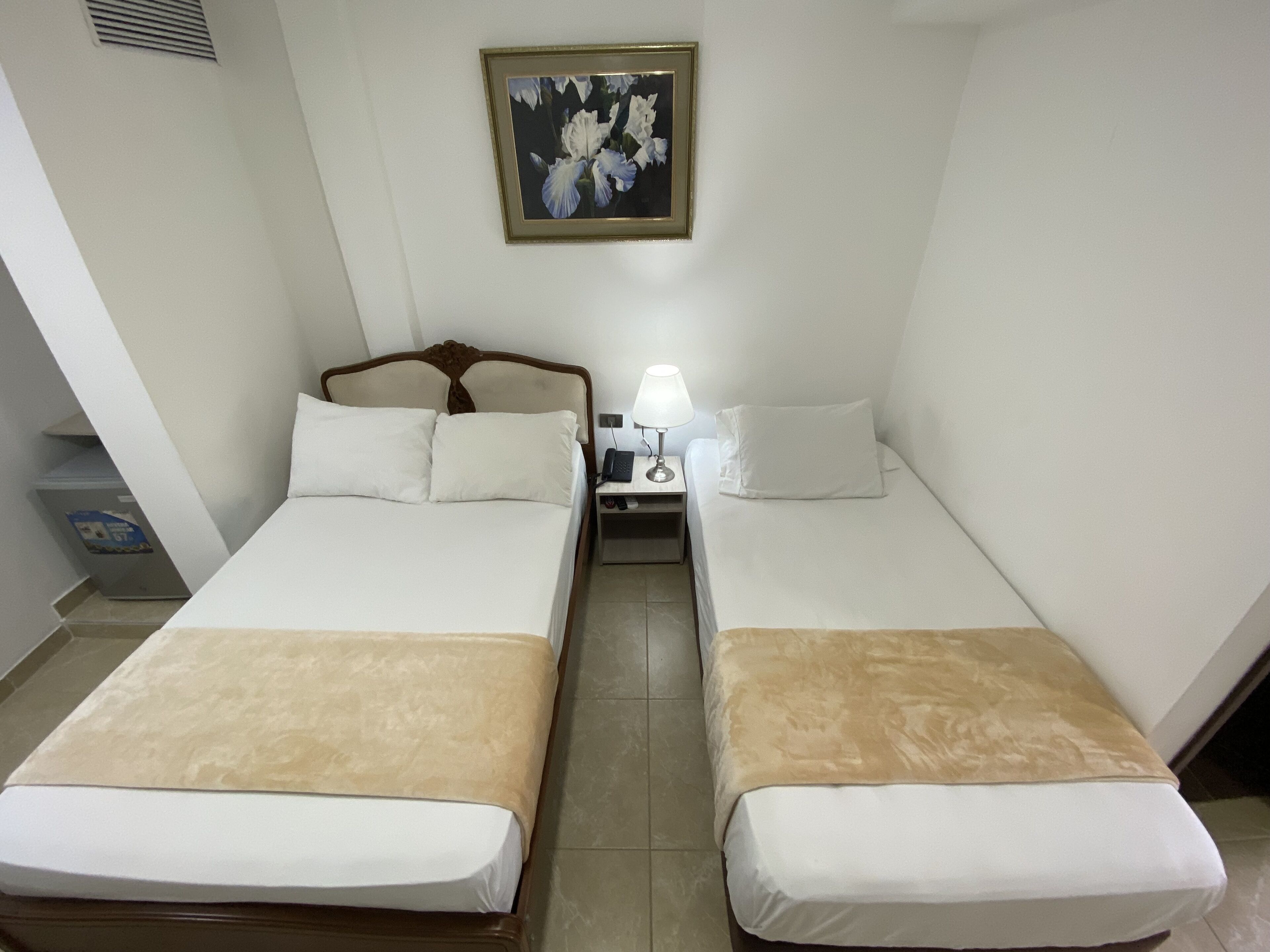 A hotel room with two single beds, a nightstand with a lamp, a framed picture on the wall, and a small trash bin.