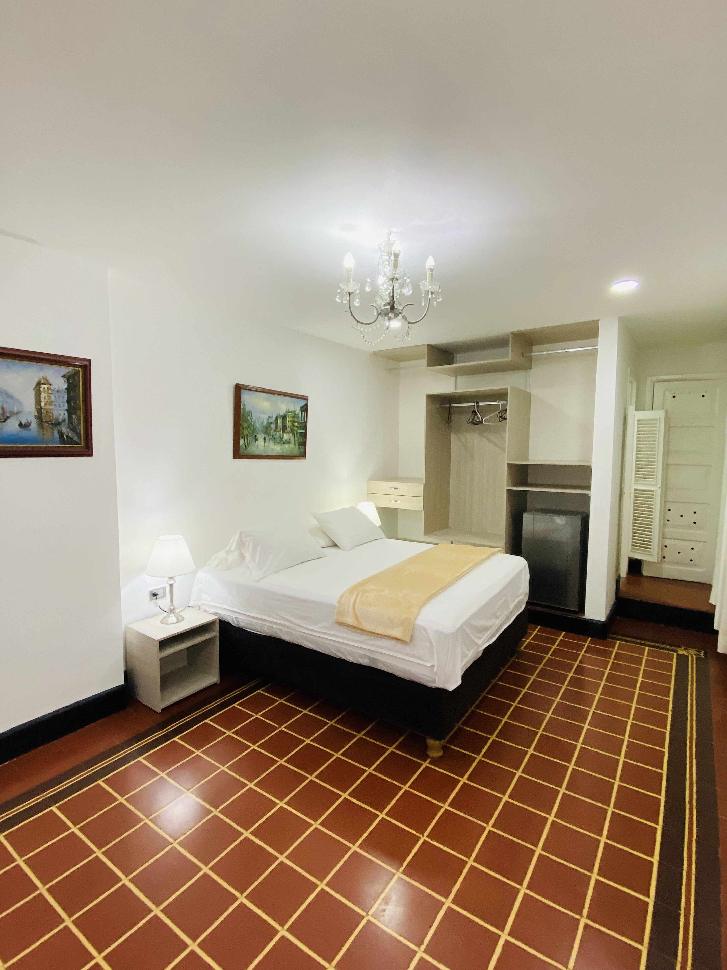 Habitación de hotel con cama, mesita de noche, lámpara, candelabro, un cuadro, armario y zona de ducha.