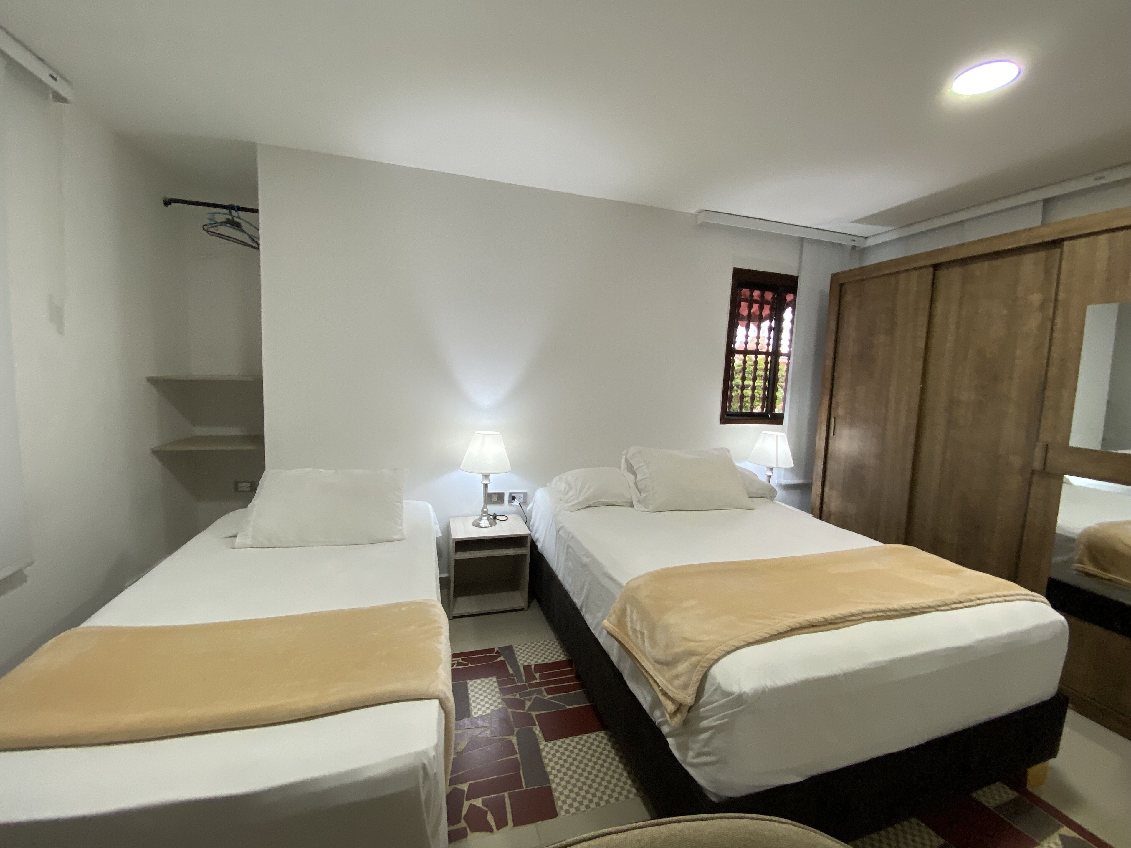 Une chambre d’hôtel avec deux lits simples, une armoire en bois, une petite table avec une lampe et une fenêtre donnant sur un paysage verdoyant.