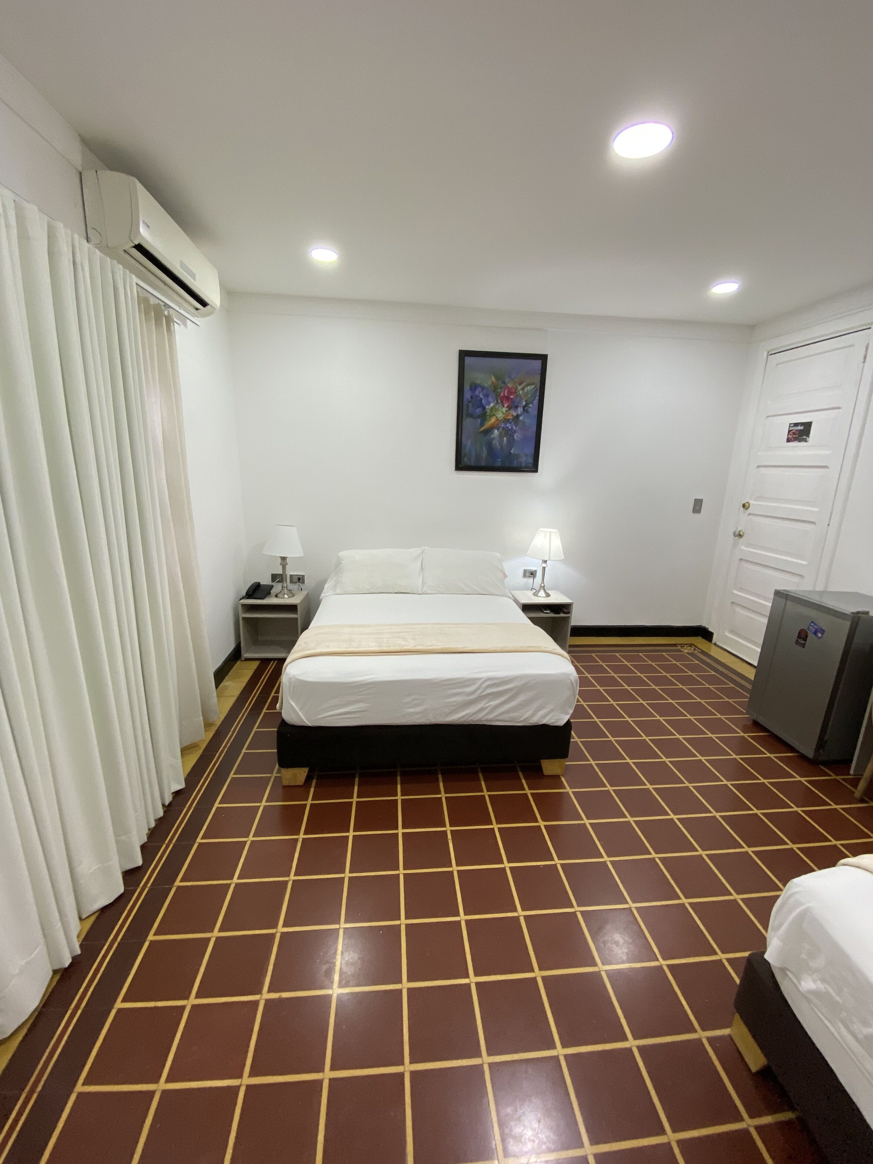 A hotel room with a bed, bedside tables, a painting, an air conditioning unit, a wardrobe, and a tiled floor.