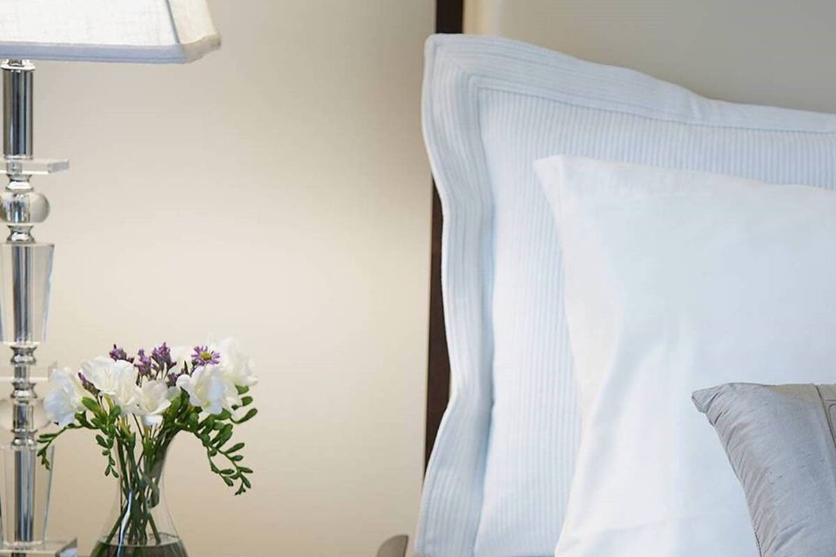 A neatly made bed with white linens, a bedside table with a lamp and a vase of flowers, and a pillow.