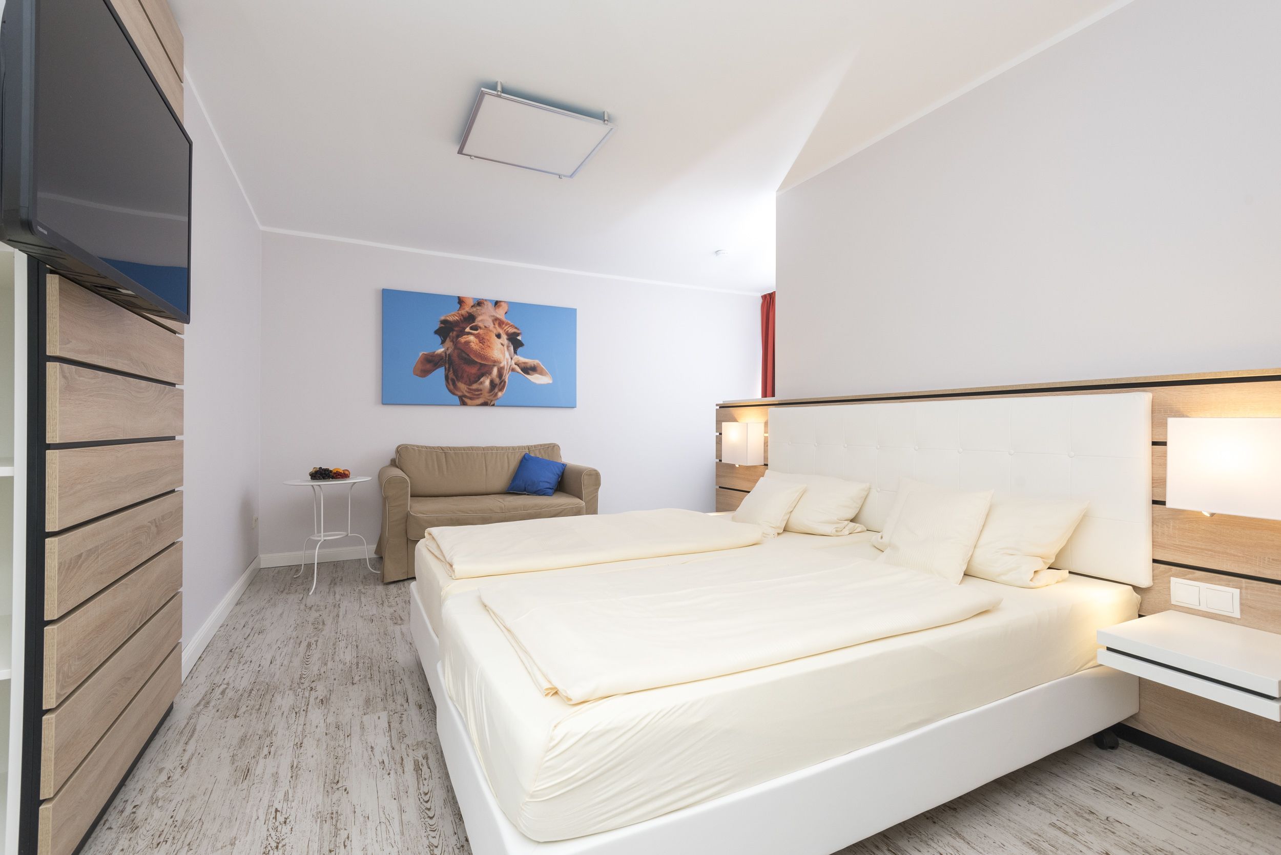 A modern hotel room with a large bed, a small table, a sofa, and a giraffe painting on the wall.