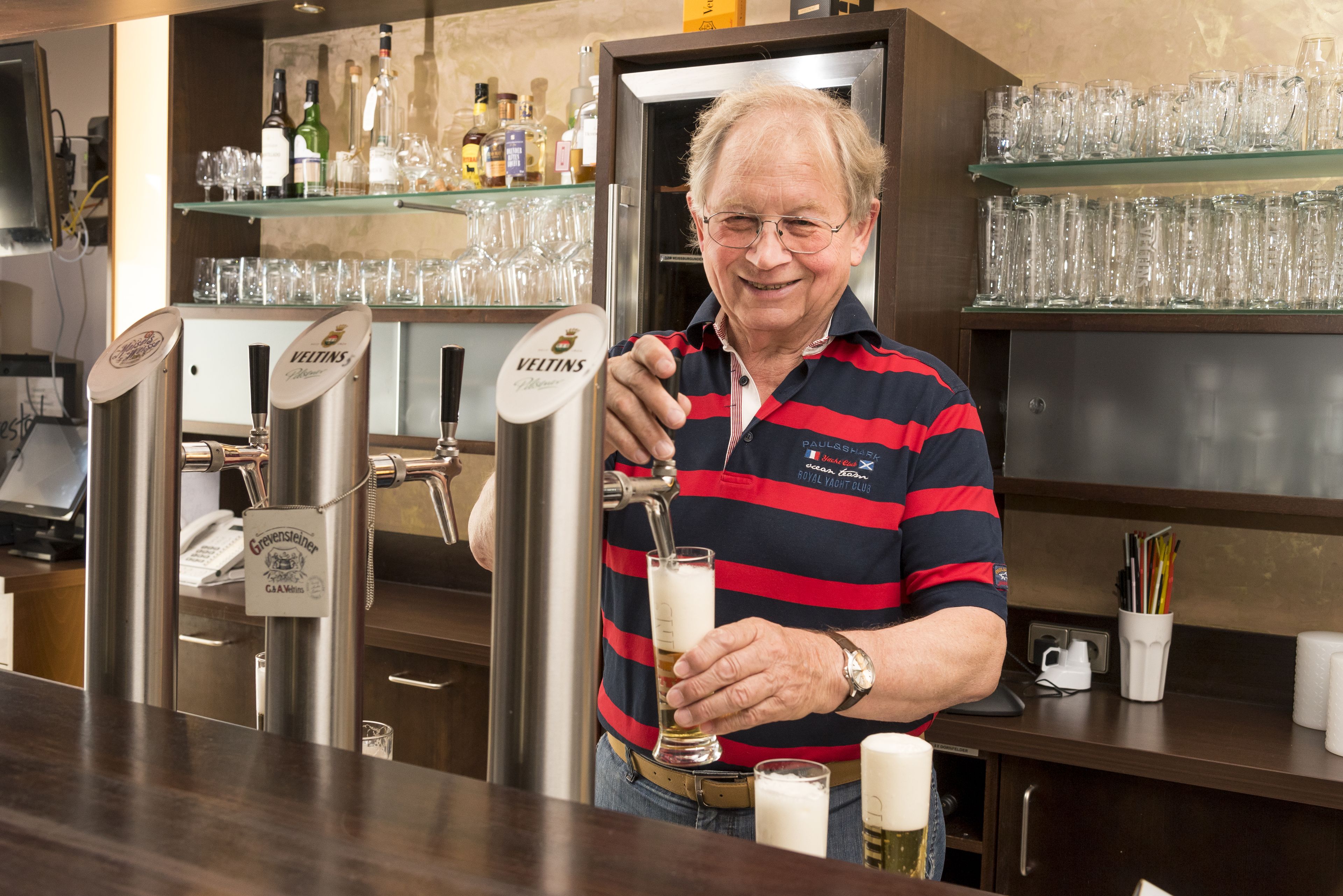 Eine Bar mit Zapfhahn, an dem ein Mann ein Getränk zapft, Regalen voller Flaschen und Gläsern sowie einer Theke mit Registrierkasse.