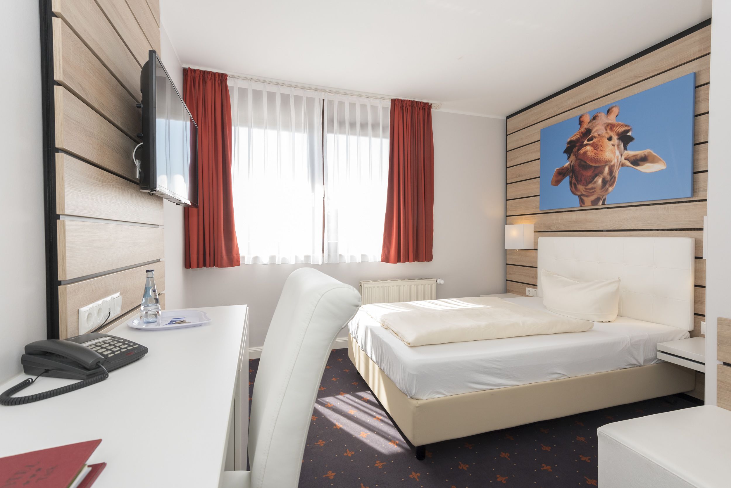 A hotel room with a bed, a desk with a telephone, a chair, a radiator, and a painting of a giraffe.