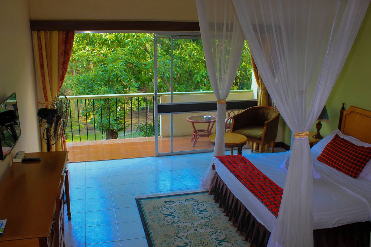 Quarto com cama, mesa e cadeira. Possui varanda com vista para área verde.