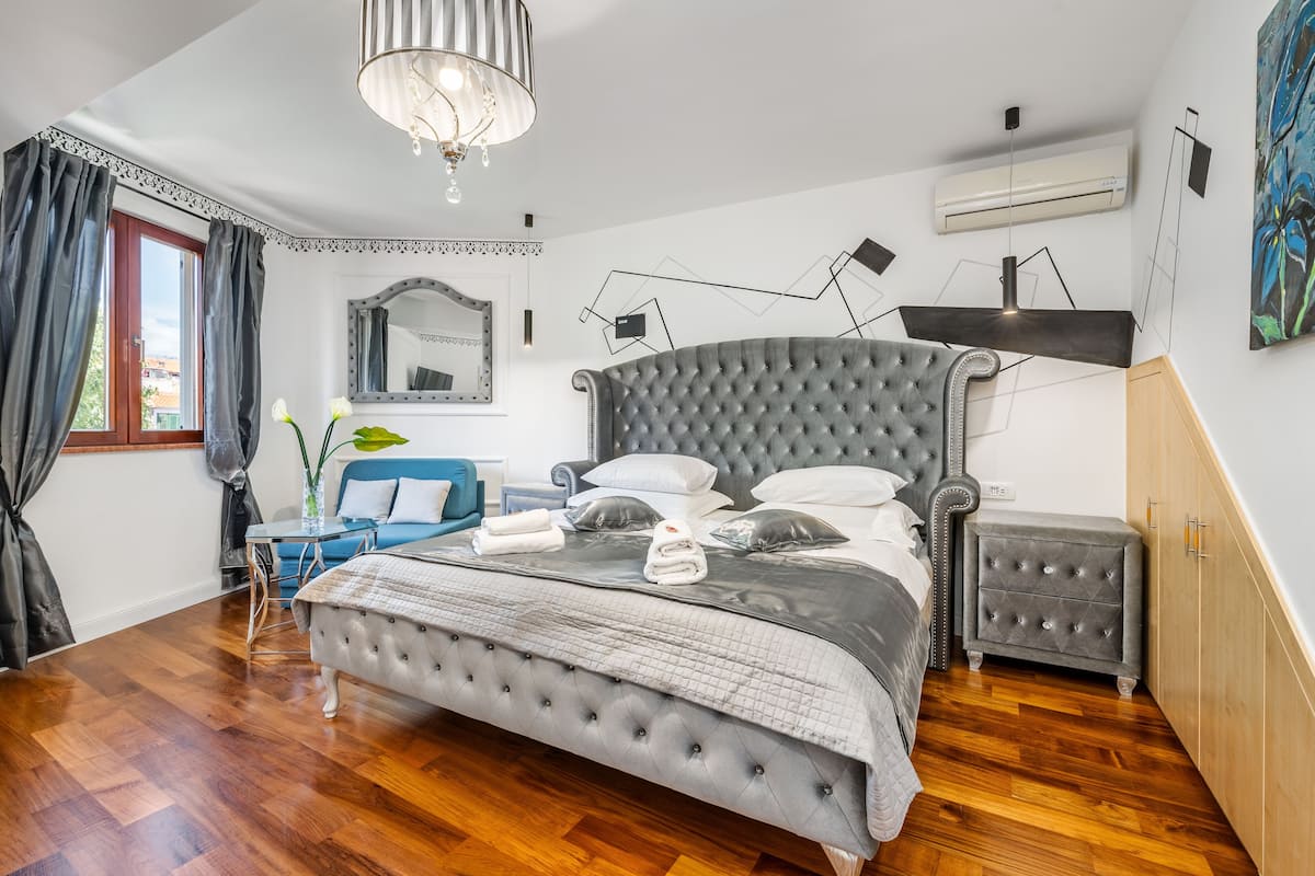 A bedroom with a large bed, a bedside table, a dresser, a mirror, and a chandelier.
