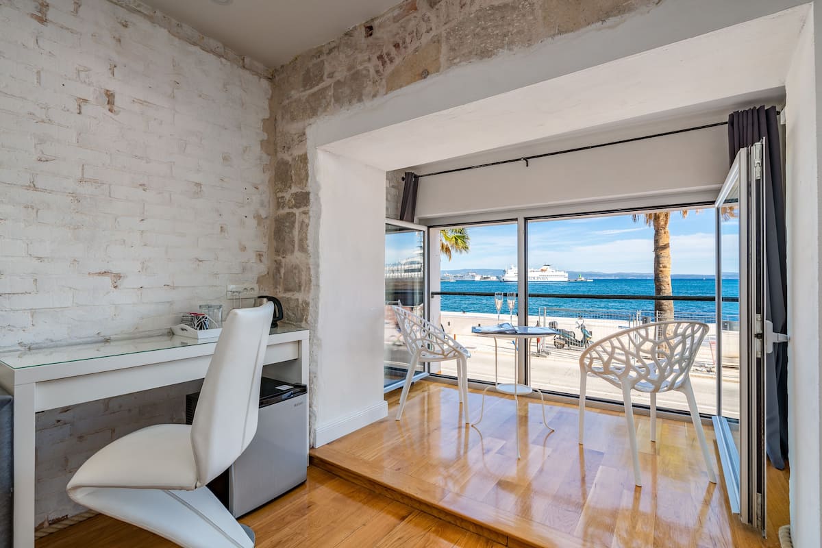 A modern room with a white brick wall, a glass desk, a white chair, and a balcony with a view of the sea and a palm tree.