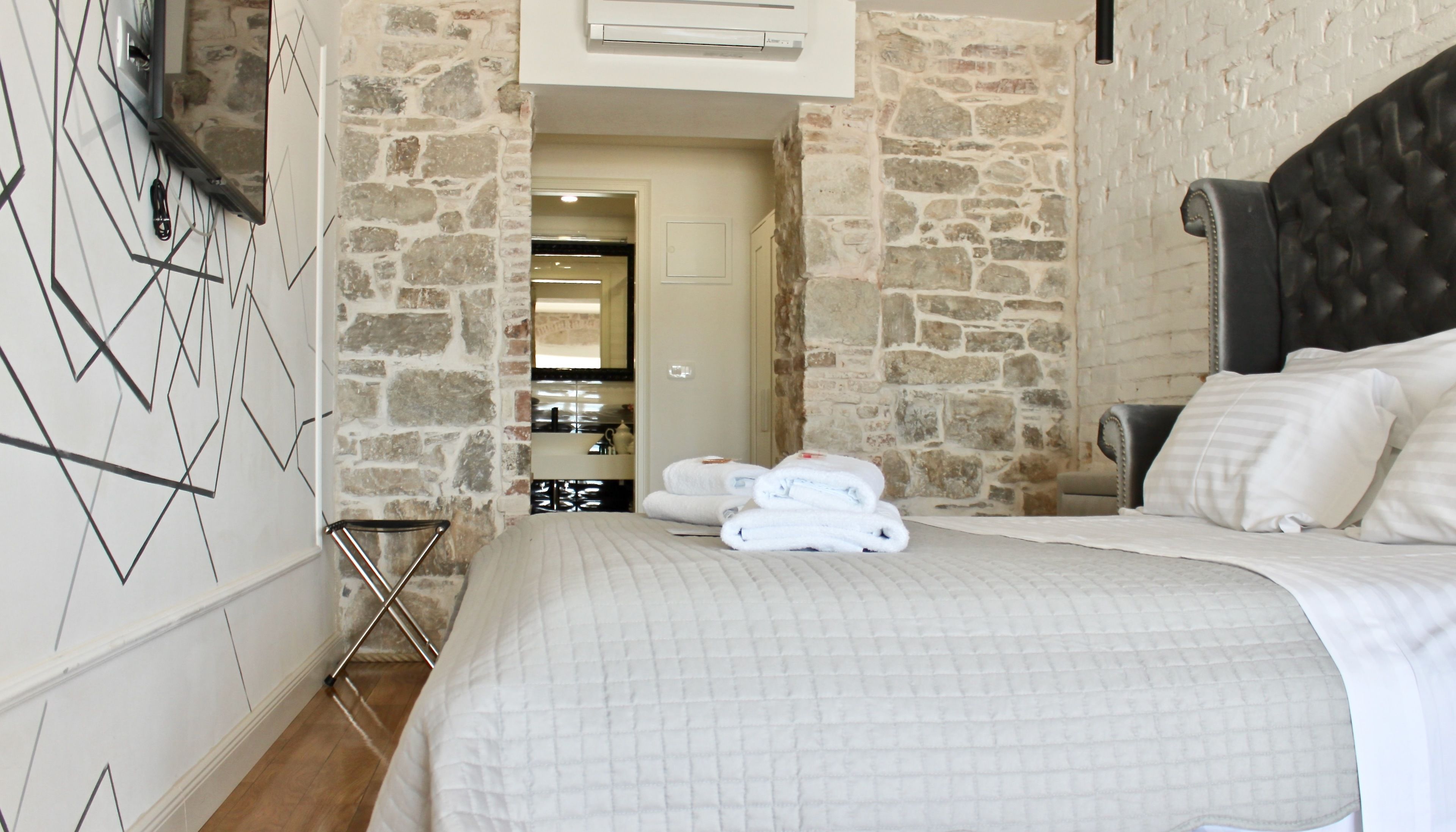 Habitació d'hotel amb llit, capçalera i paret de pedra. Hi ha una porta que dona a un bany i un passadís.