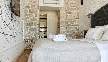 A hotel room with a bed, a headboard, and a stone wall. There is a door leading to a bathroom and a hallway.