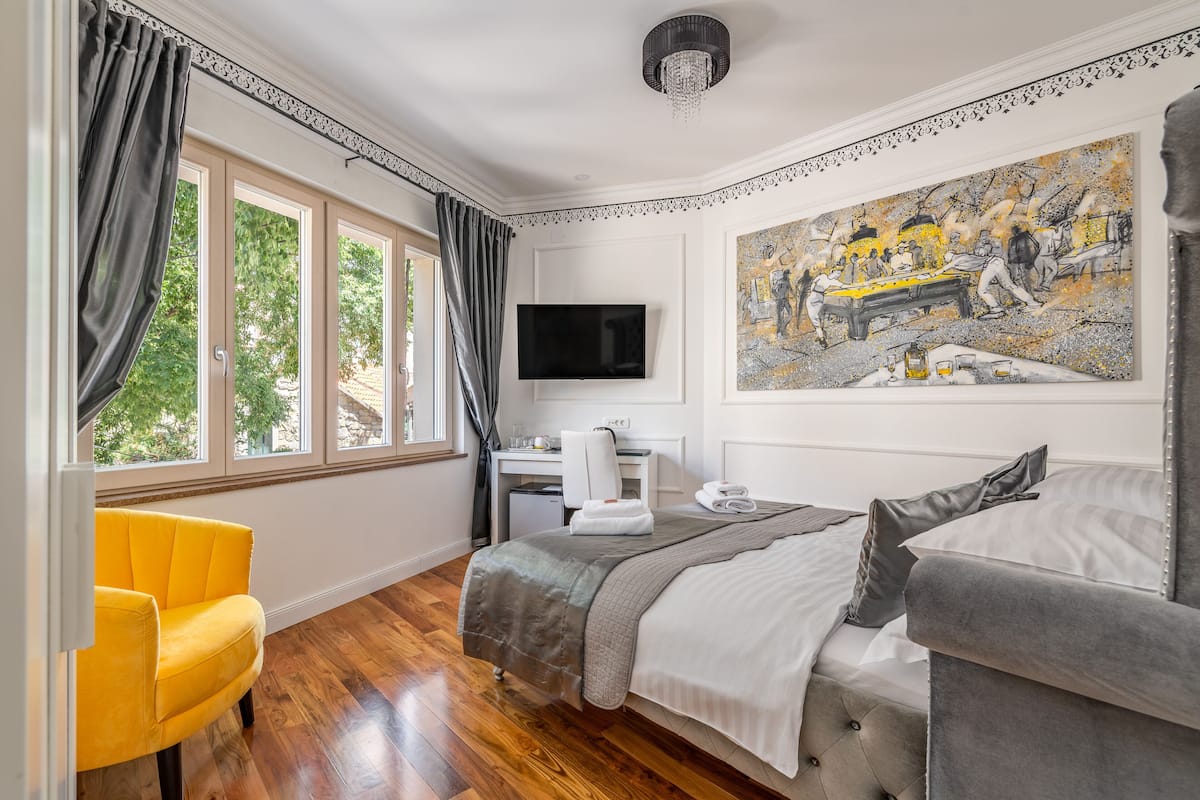 A bedroom with a bed, a desk, a chair, a painting, and a television.