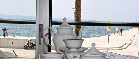Una mesa con un juego de té y una computadora portátil, con vistas a un muelle con una palmera y gente caminando.