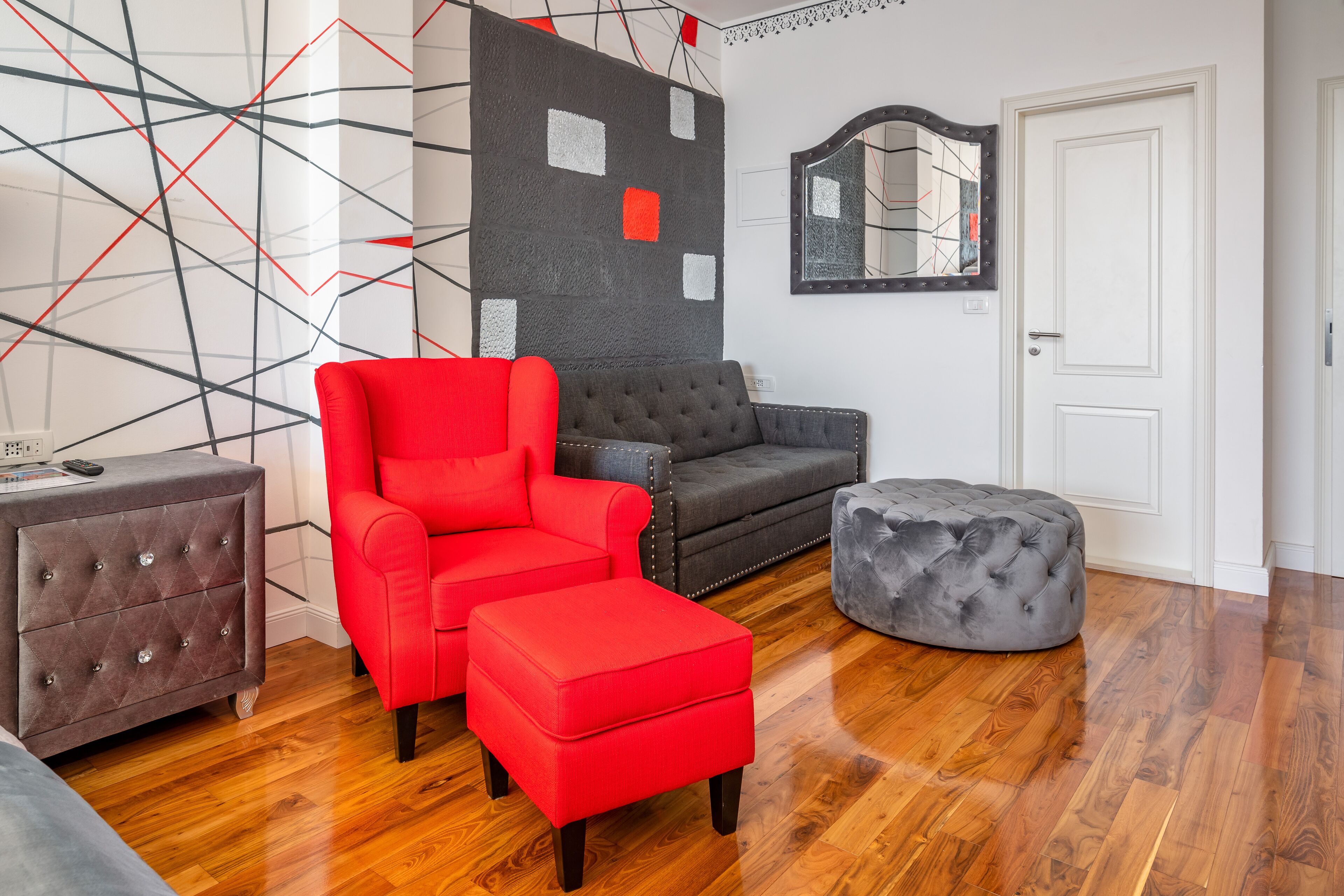 A modern living room with a red armchair, grey sofa, and ottoman, wooden flooring, and a geometric wall design.
