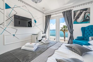 A hotel room with a bed, a desk, a TV, a balcony with a view of the sea, and a painting.