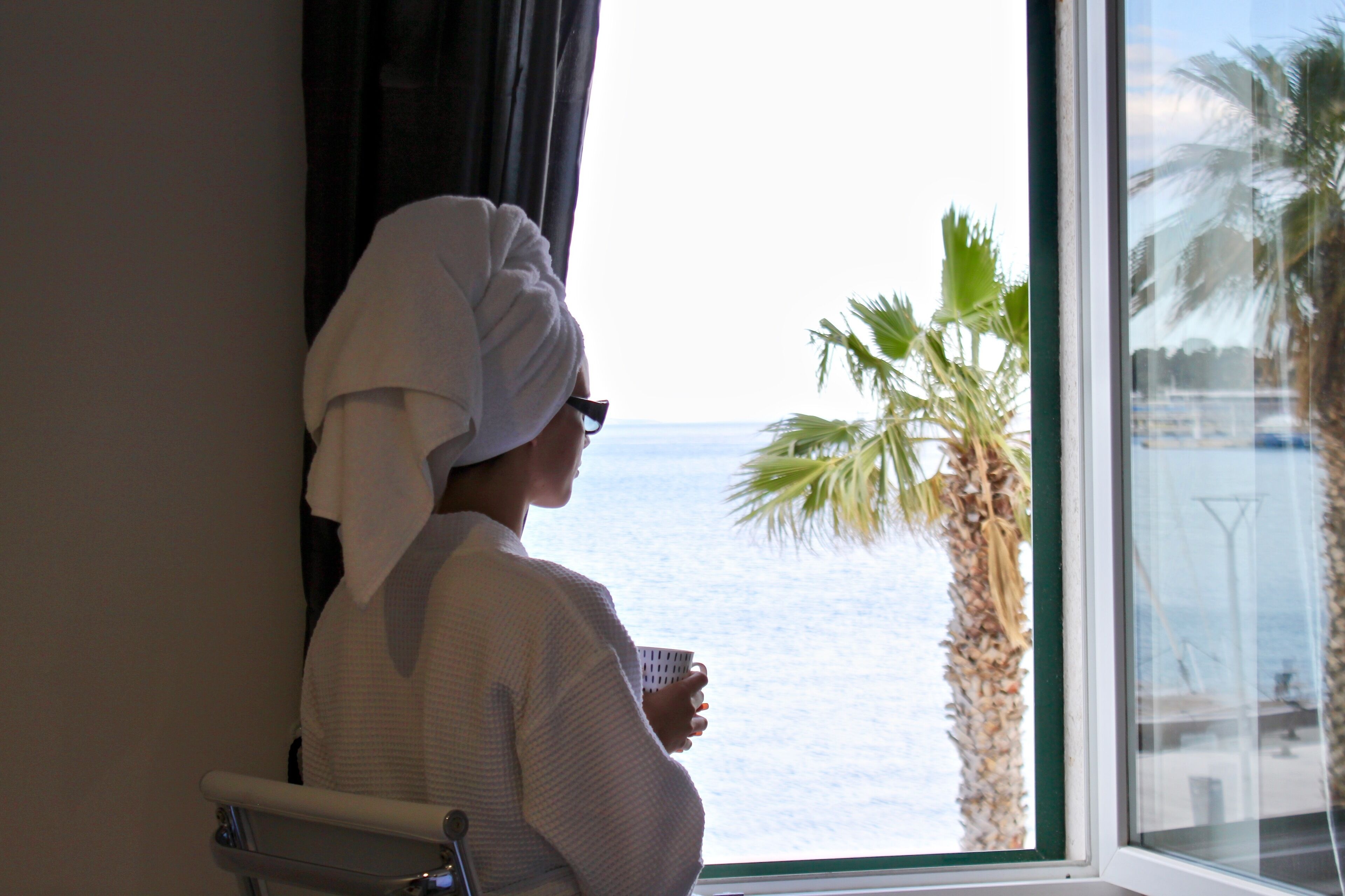 A person in a white robe with a towel on their head, holding a cup, standing by a window with a view of the sea and palm trees.