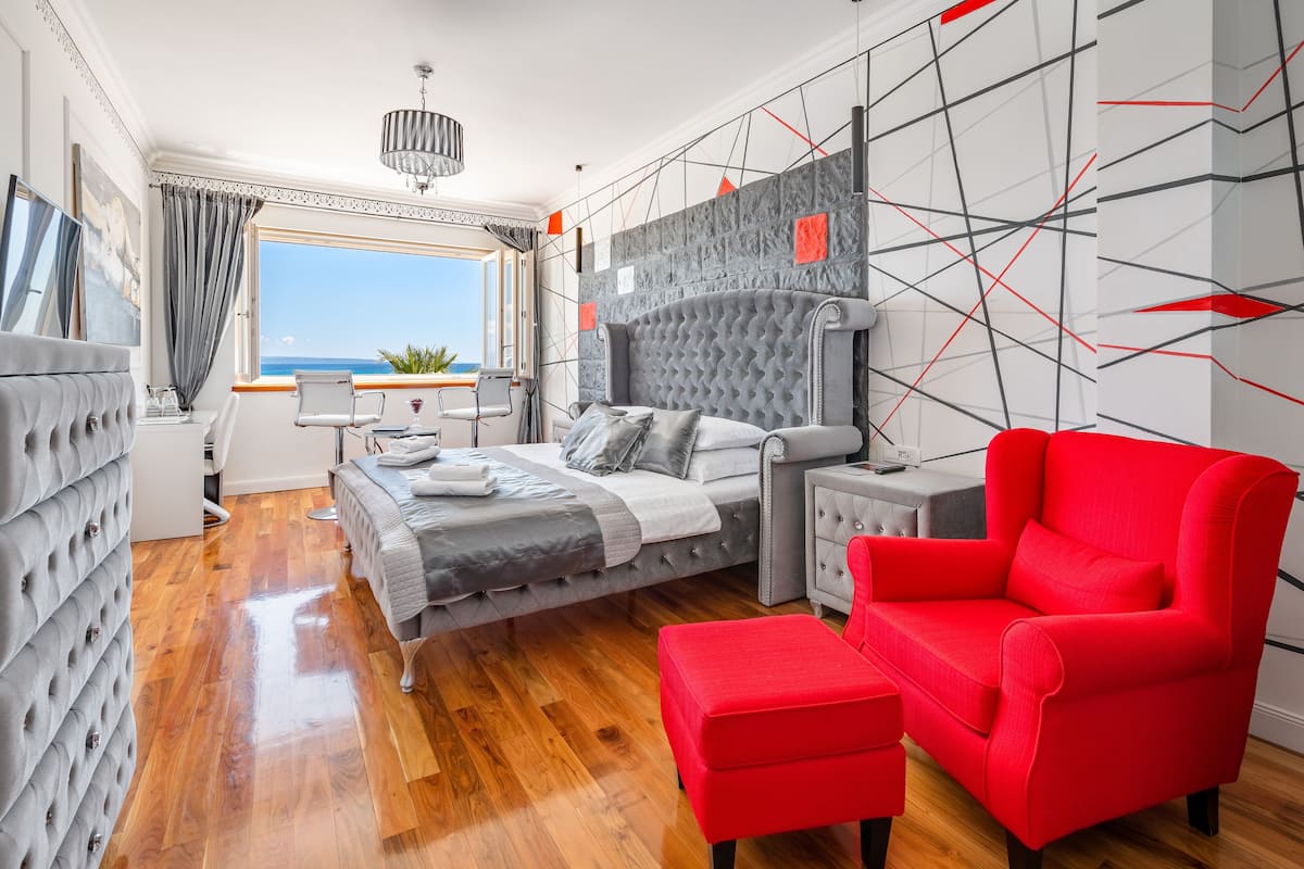 A bedroom with a bed, a red armchair, a nightstand, and a view of the ocean.
