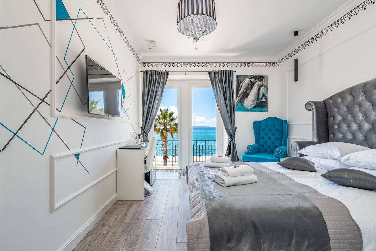 A modern bedroom with a large bed, a blue armchair, and a view of the sea.