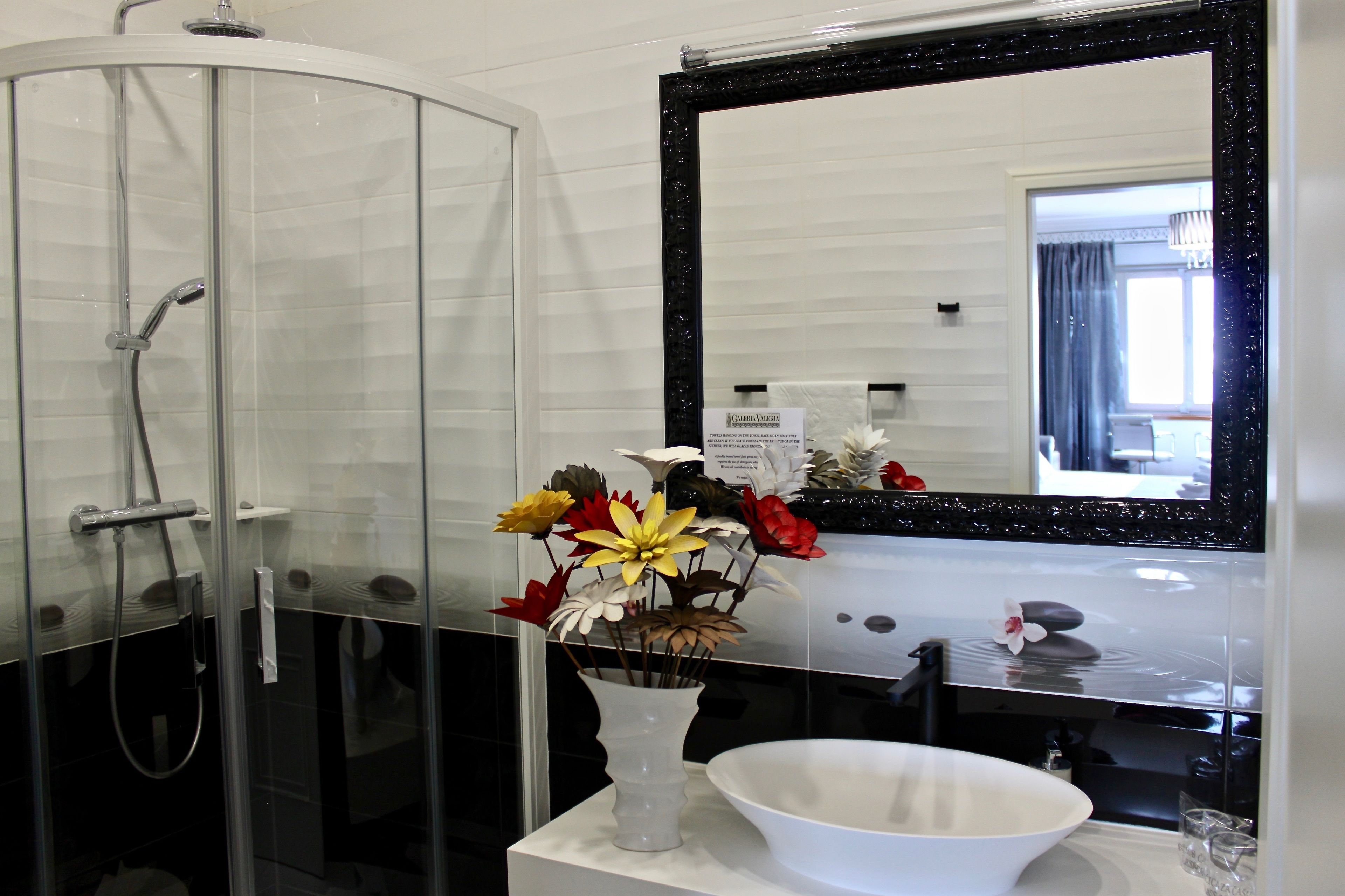 Un baño con ducha de vidrio, un lavabo blanco, un espejo con marco negro y un jarrón con flores artificiales.
