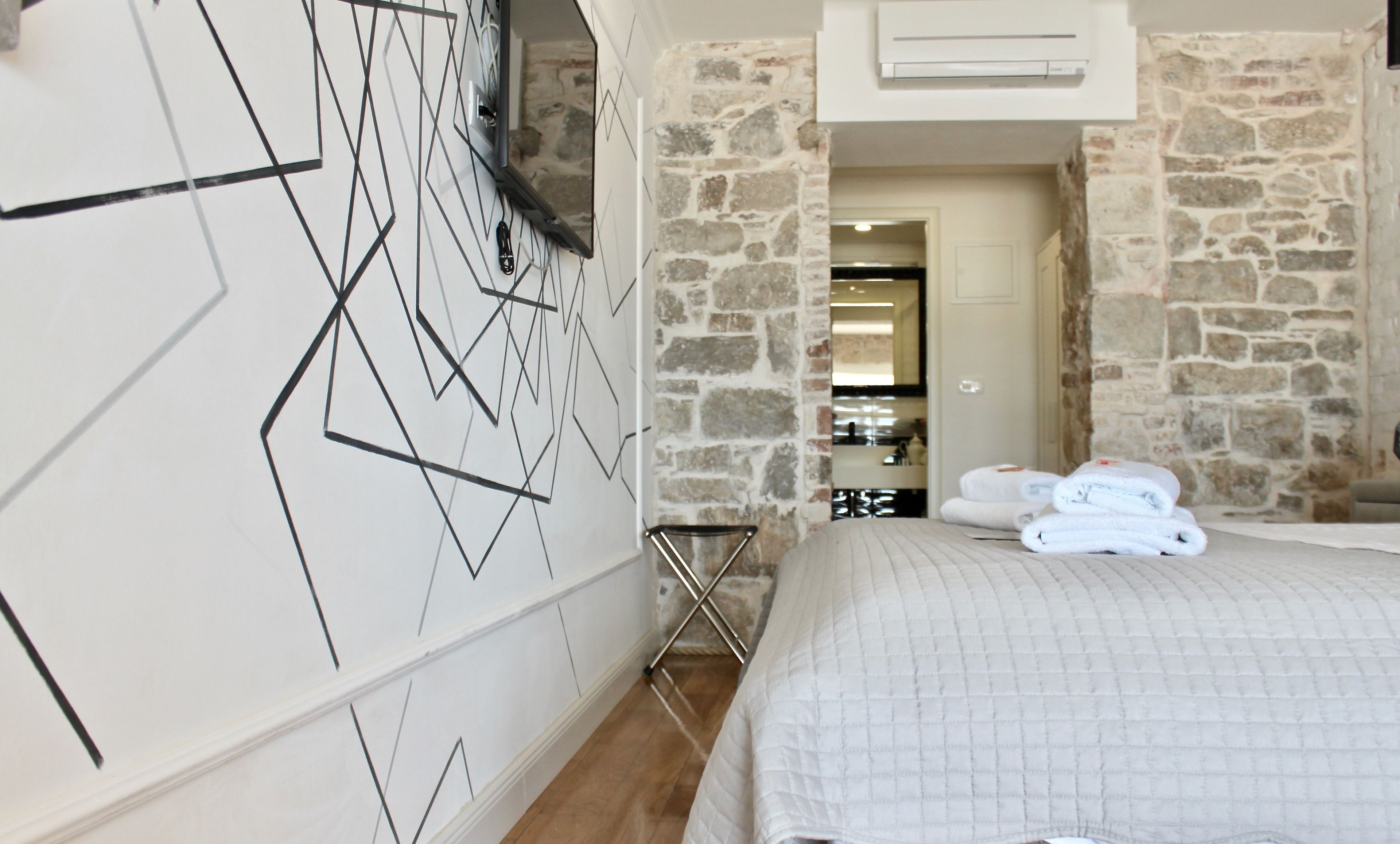 A bedroom with a bed, a stone wall, a TV mounted on the wall, and a bathroom visible through an open door.