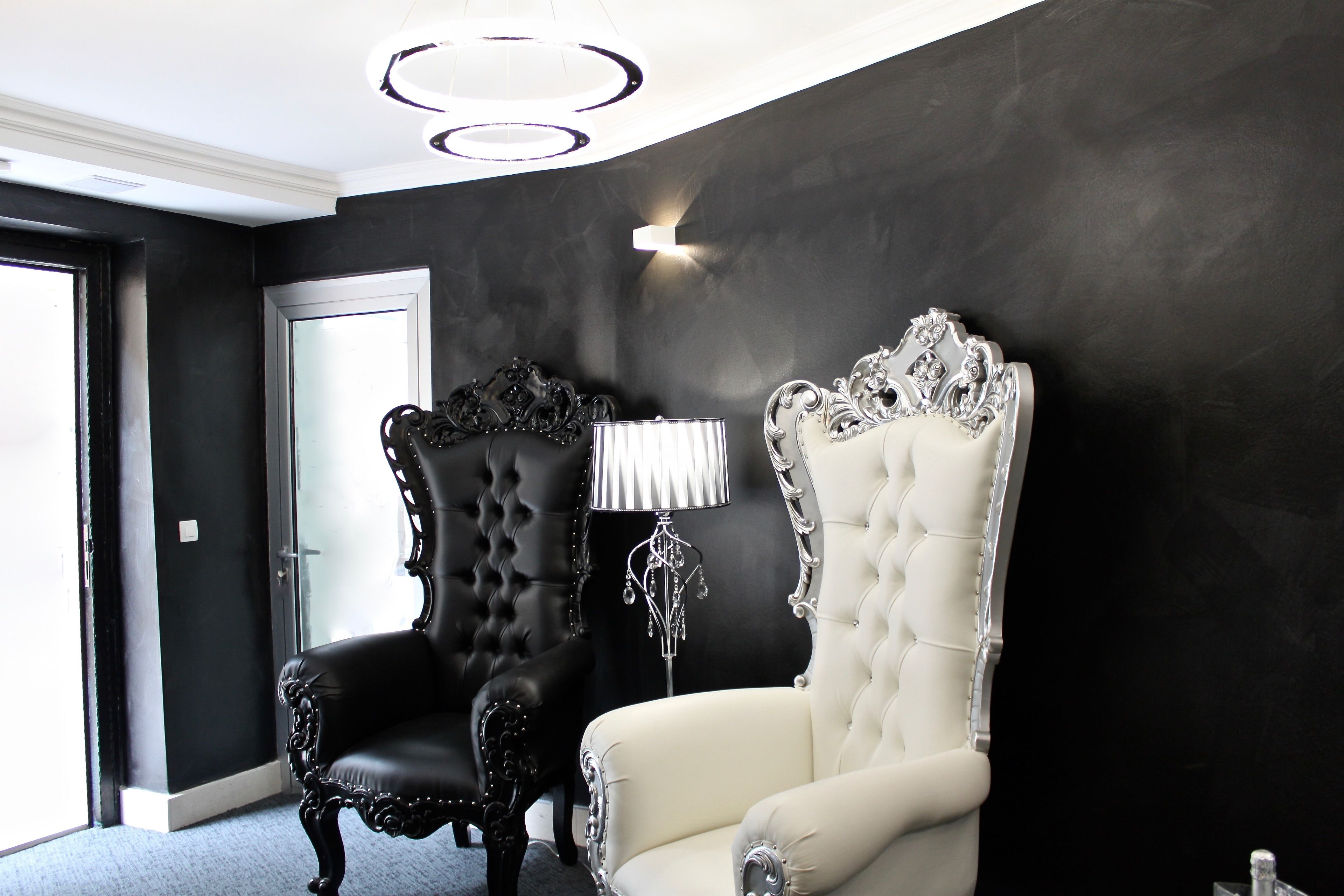 Una habitación con dos sillones decorados, una araña y una lámpara frente a una pared oscura.