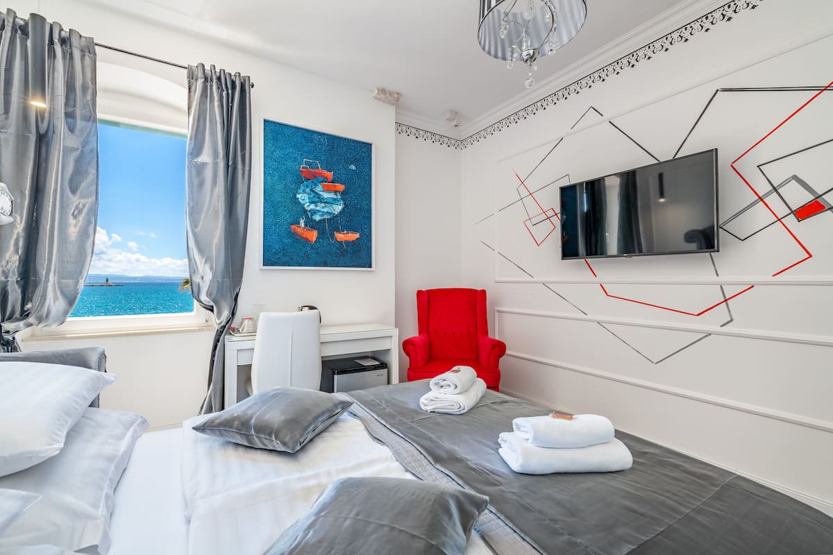 A hotel room with a bed, a desk, a red chair, a TV, and a painting of boats on the wall.