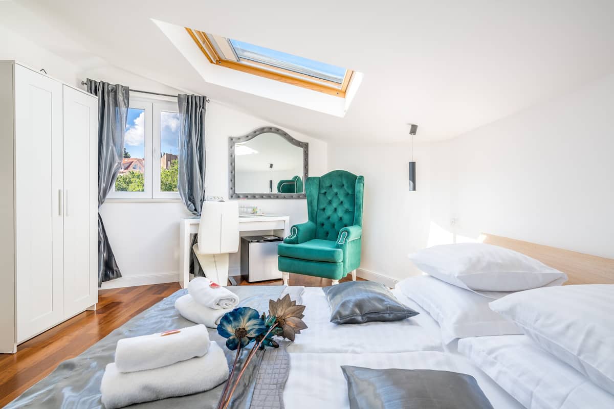 A bedroom with a skylight, a vanity with a mirror, a teal armchair, and a neatly made bed with pillows and towels.