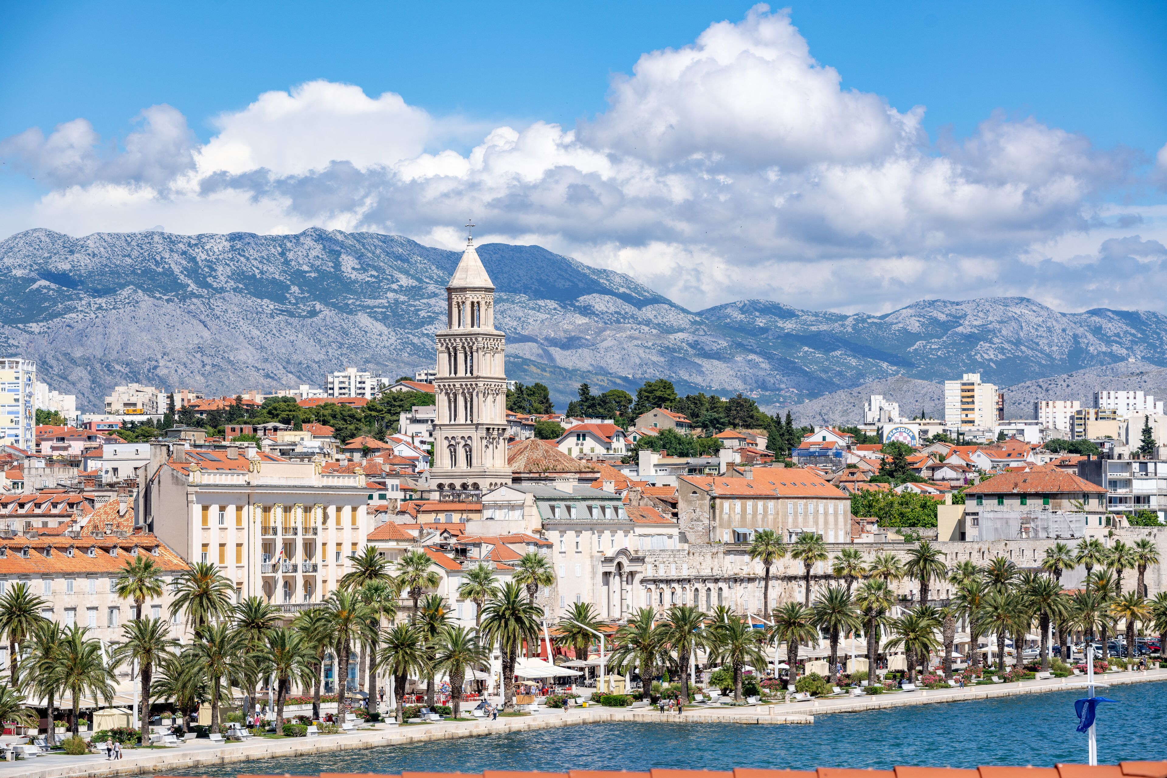 A coastal cityscape with a prominent tower, palm-lined promenade, and mountainous backdrop.