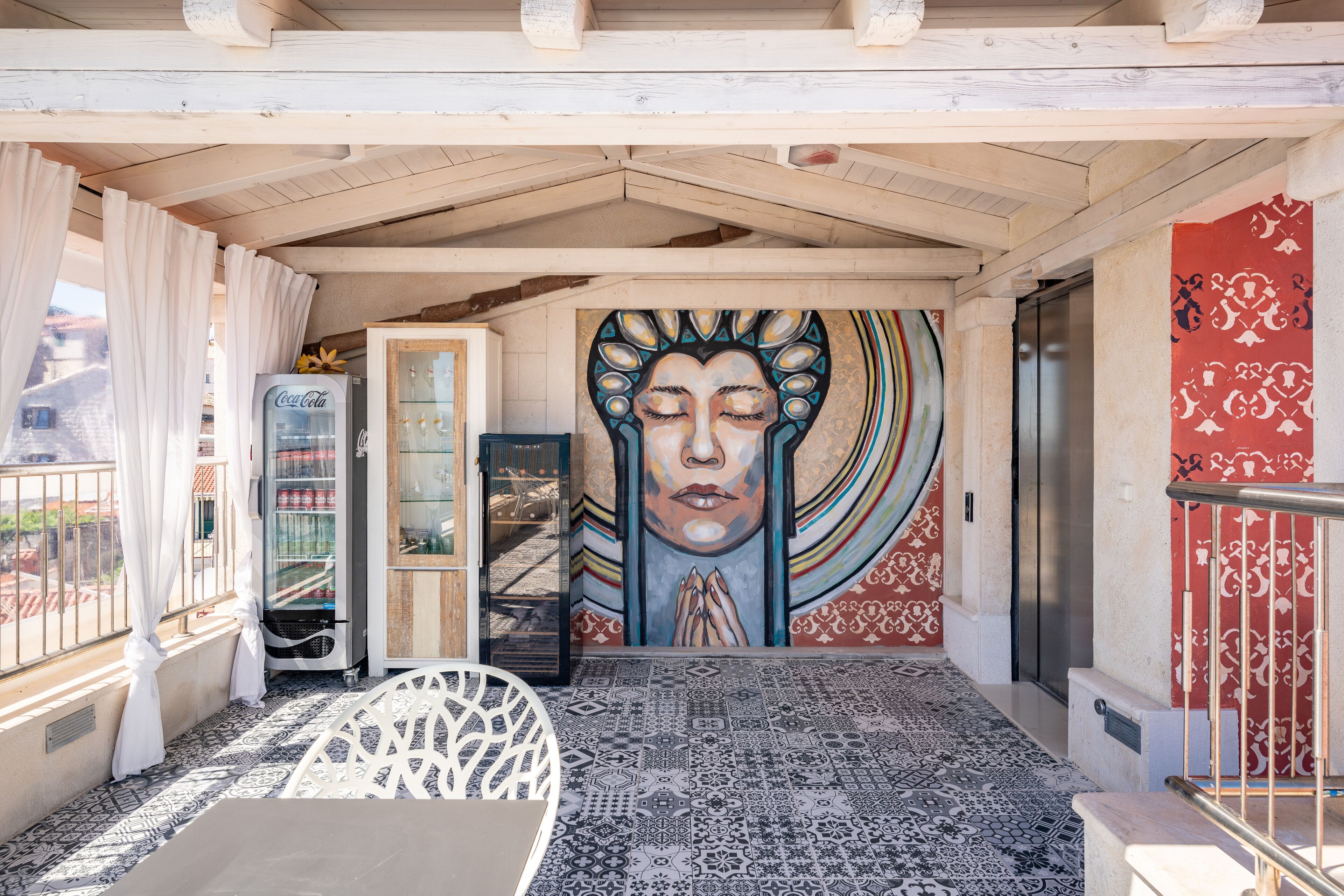 A covered outdoor area with a tiled floor, a Coca-Cola refrigerator, a glass display case, and a mural on the wall.