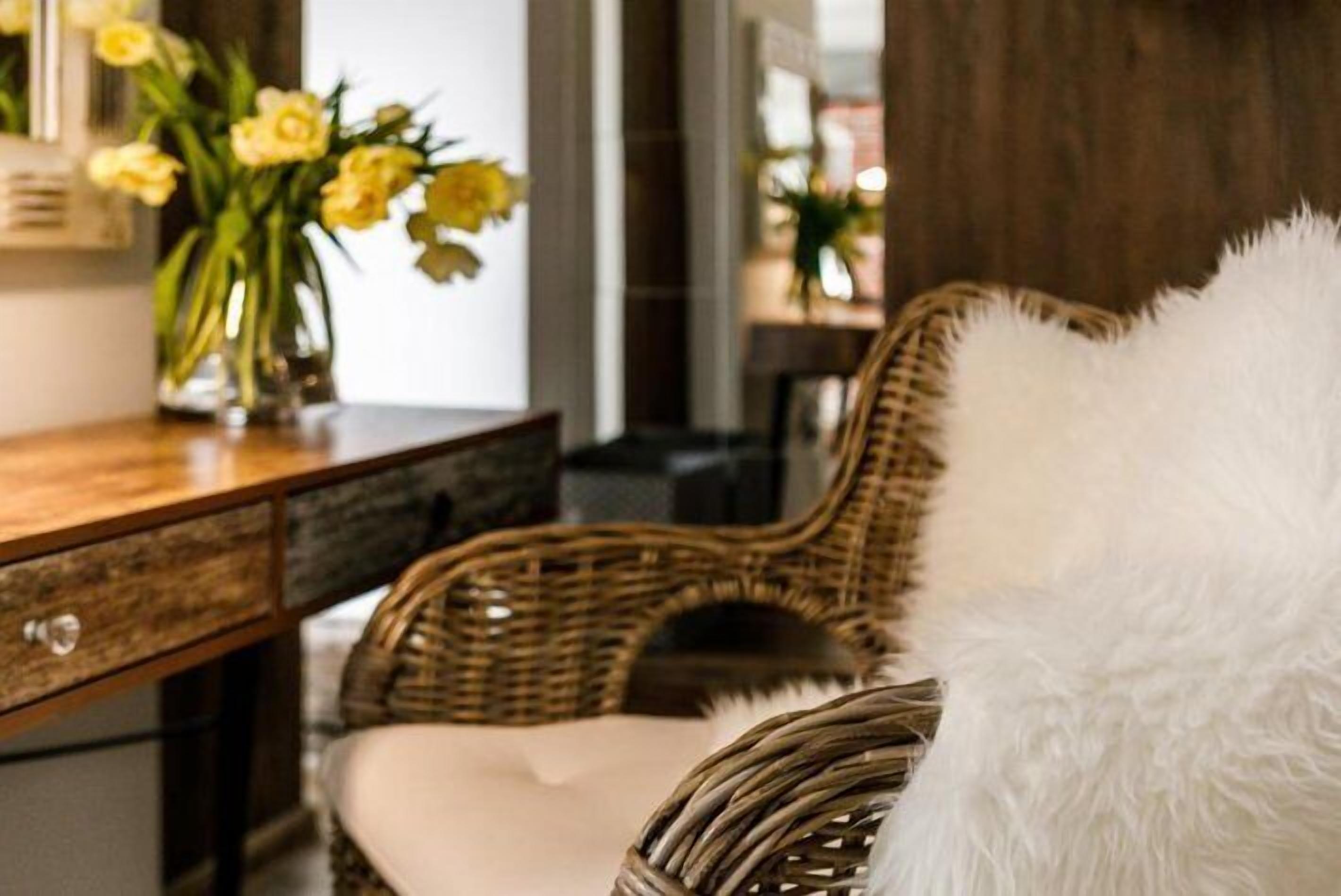Une chaise en osier avec un coussin blanc, un bureau en bois orné d’un vase de fleurs jaunes et un coussin moelleux blanc.
