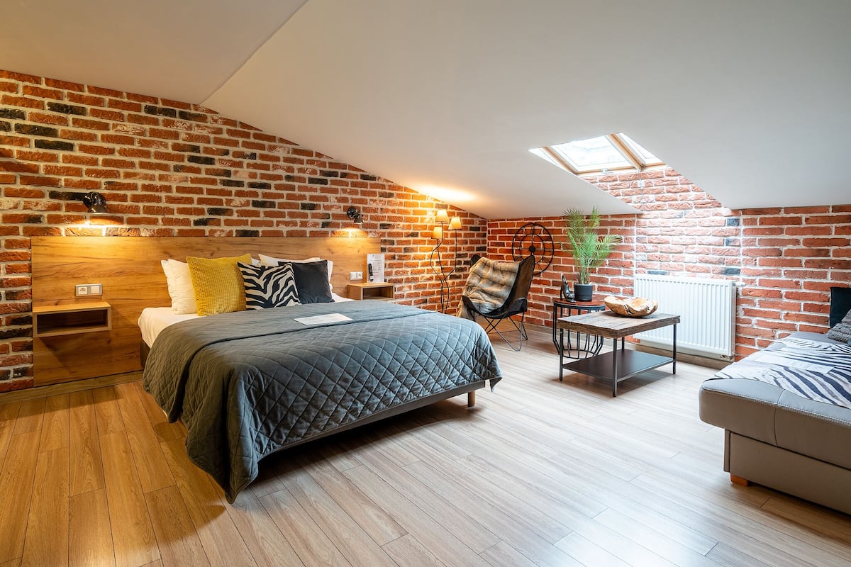 A loft bedroom with a sloped ceiling, exposed brick walls, a wooden bed, a small table, a chair, and a sofa.