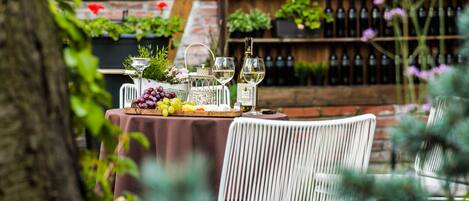 Un área de comedor al aire libre con una mesa dispuesta para dos personas, rodeada de follaje y una pared de ladrillo con botellas de vino al fondo.
