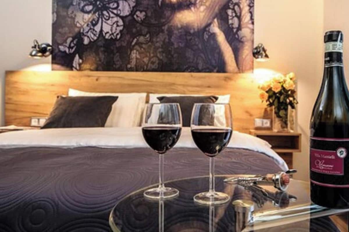 A bedroom with a bed, two wine glasses, a bottle of wine, and a painting on the wall.