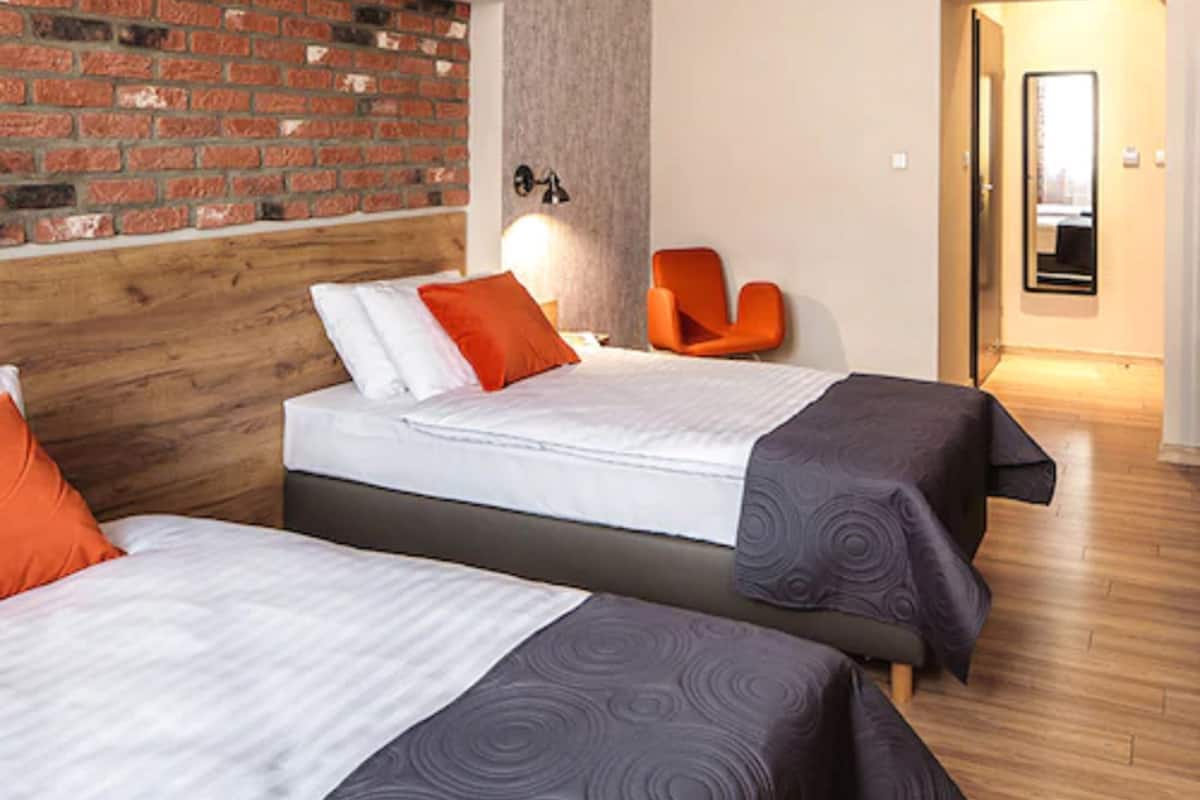 A hotel room with two beds, a brick wall, a wooden headboard, an orange chair, and a mirror.