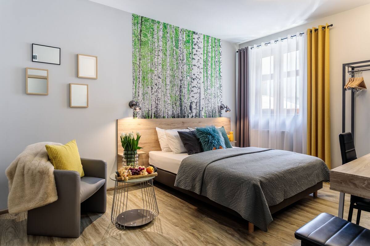 A modern hotel room with a bed, a sofa, a small table with fruit, a mirror, and a wall art of trees.