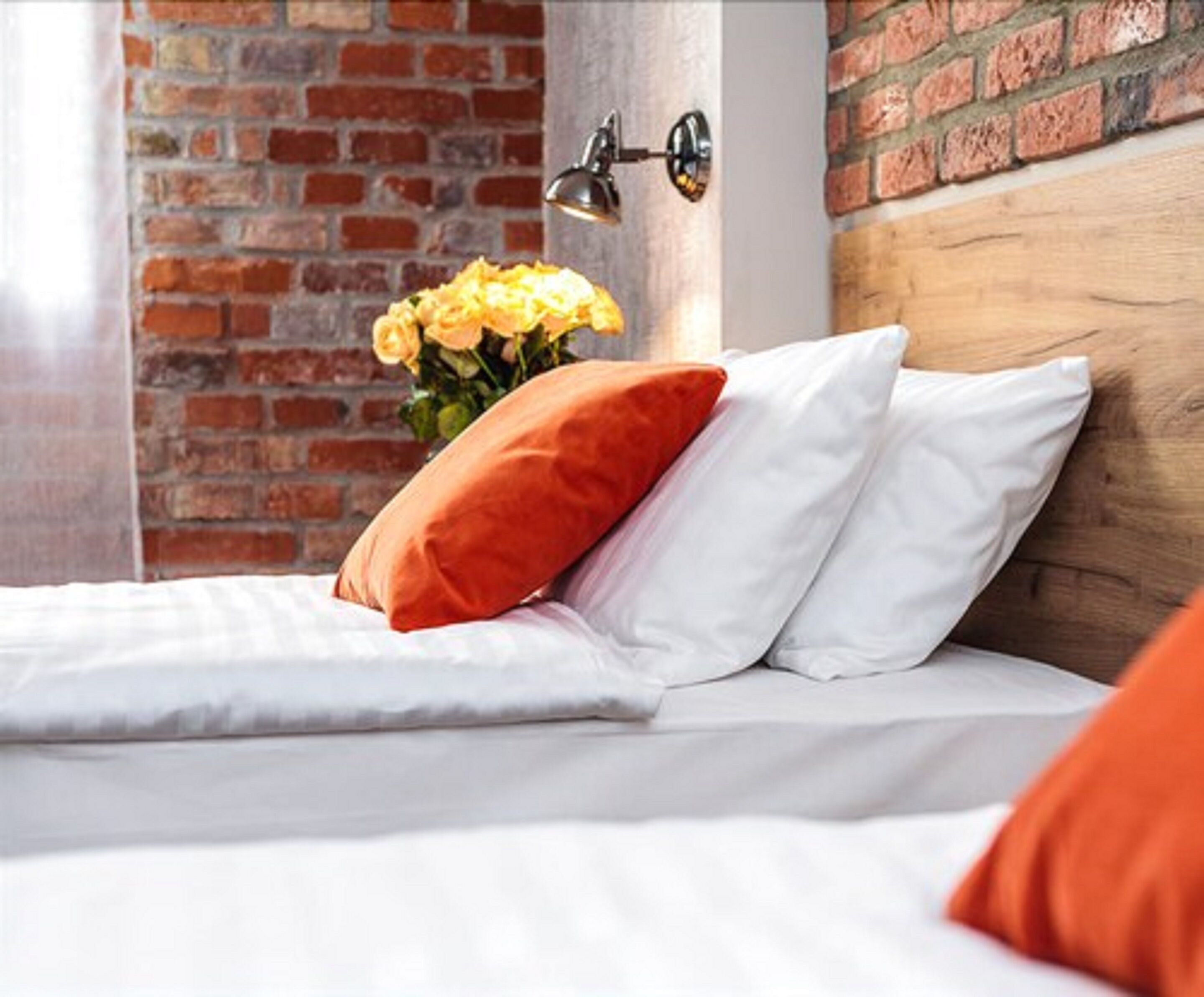 A neatly made bed with white and orange pillows, a brick wall, and a wall-mounted lamp.