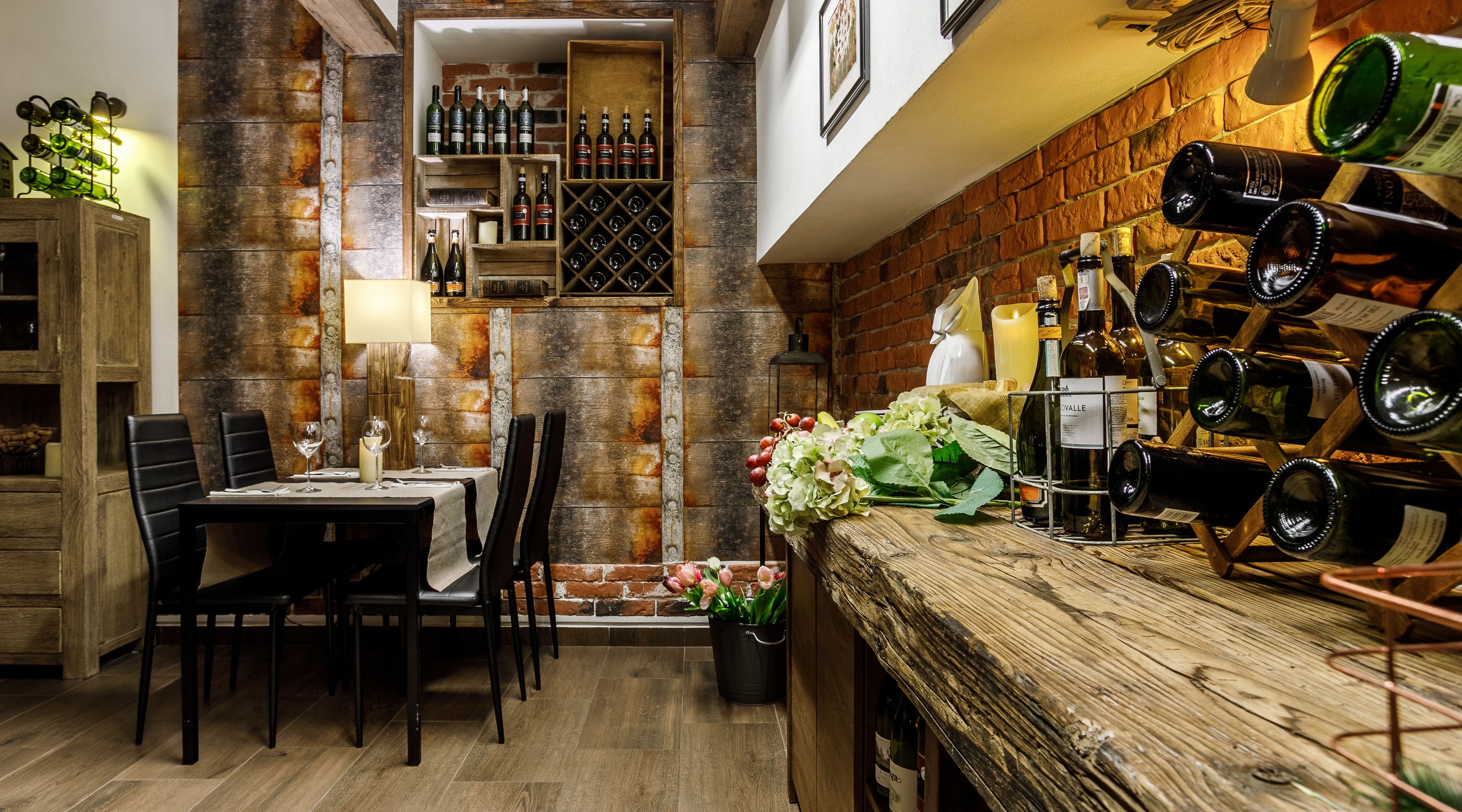 Un comedor acogedor con pared de ladrillo, mesa y sillas de madera. Hay botellas y copas de vino en estanterías y una planta en maceta sobre la mesa.