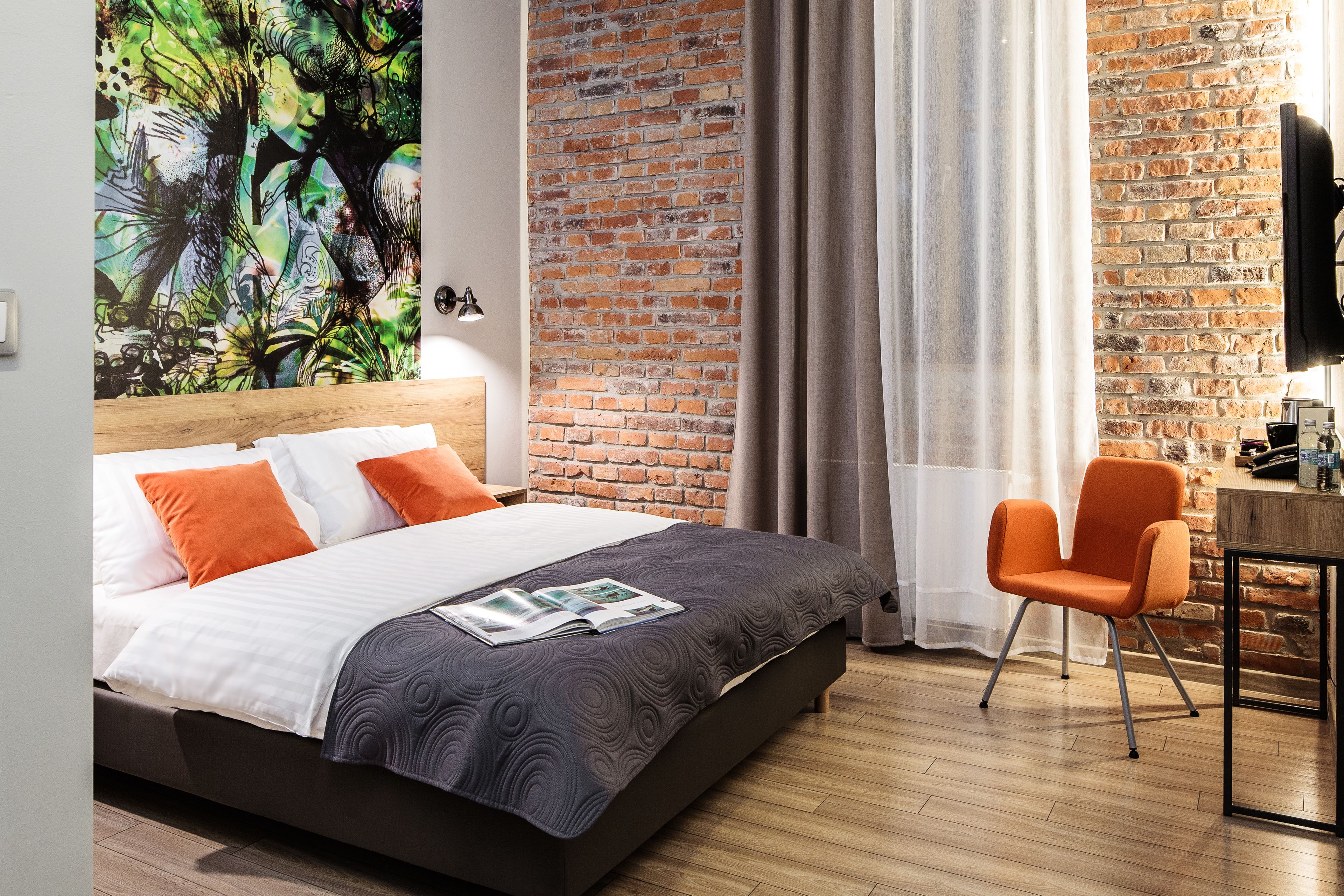 A modern hotel room with a brick wall, a wooden bed, an orange chair, and a flat-screen TV.