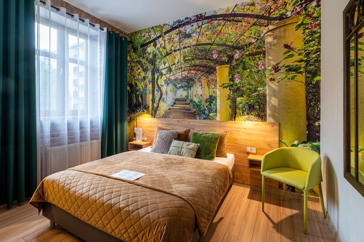 A hotel room with a large bed, a green chair, a mural of a garden, and a wooden headboard.