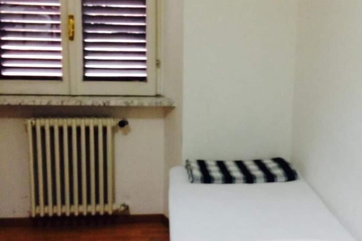 Une petite chambre avec un lit simple, un radiateur et une fenêtre équipée de stores.