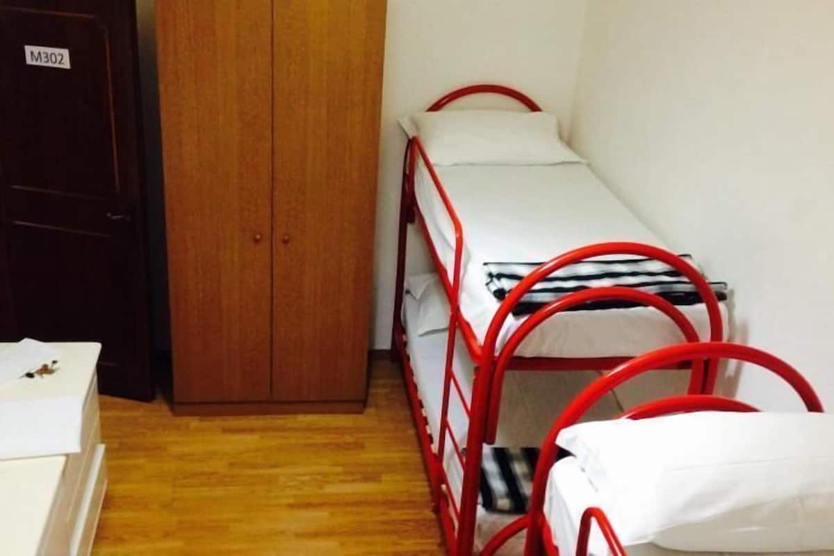 Une petite pièce étroite, avec une armoire en bois, un lit simple doté d’un cadre en métal rouge et une table de chevet blanche.